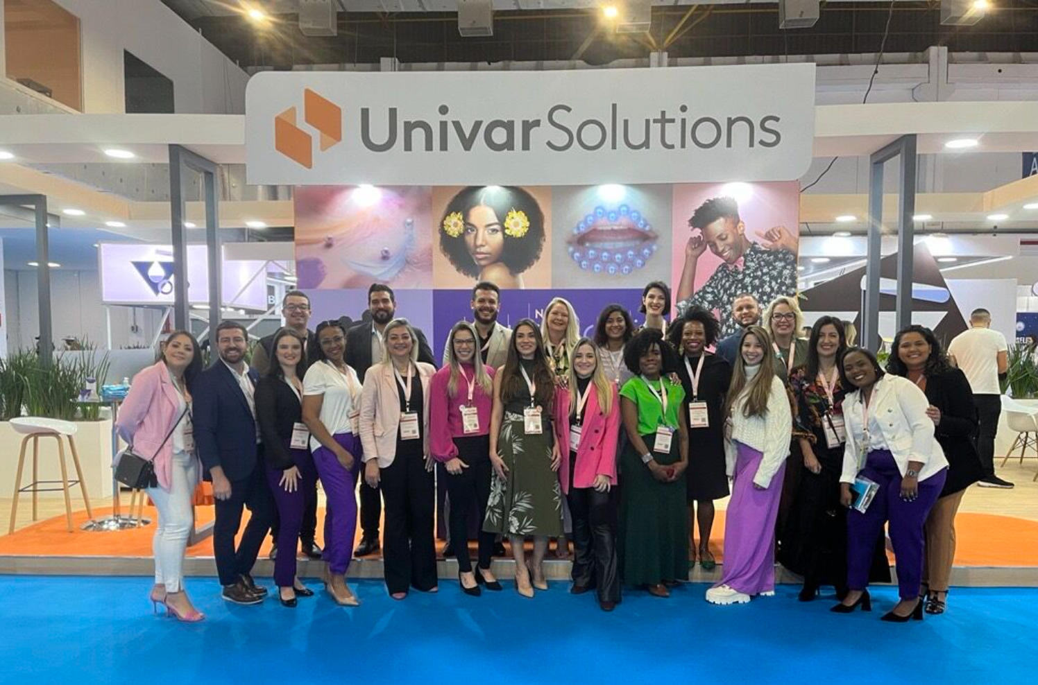 Group of diverse professionals posing at a conference booth with a 'Univar Solutions' sign, featuring colorful artwork and portraits in the background.