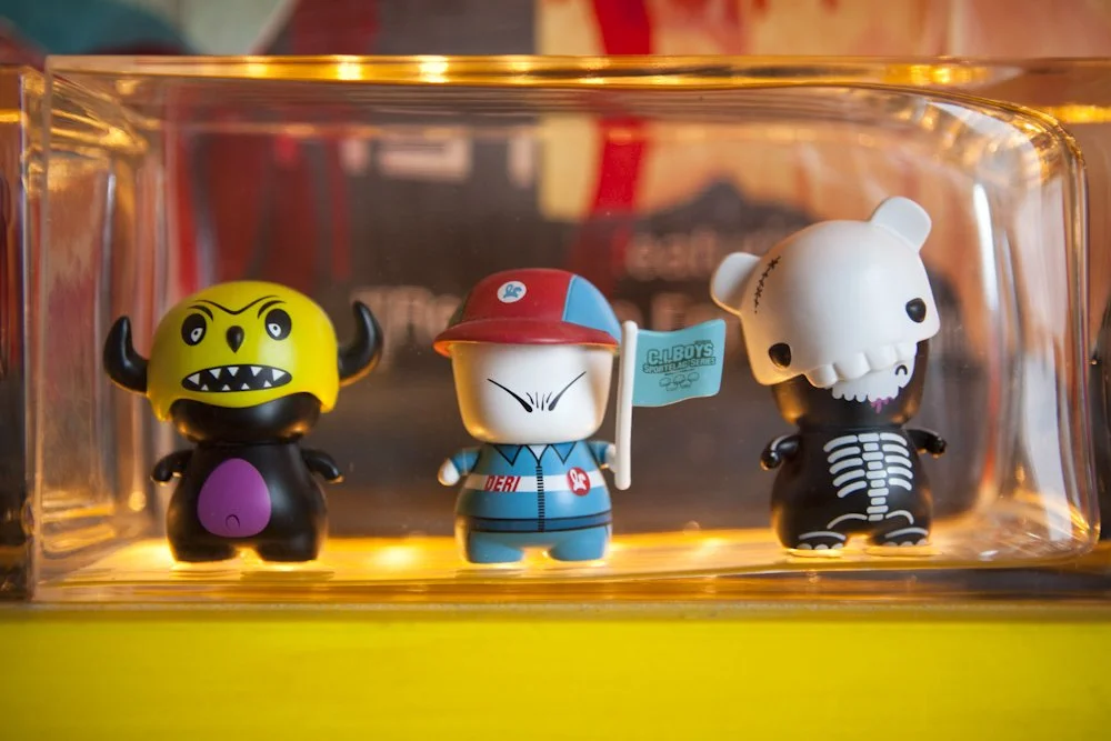 Three small collectible figures inside a clear plastic box: a black and yellow monster with horns and sharp teeth on the left, a cartoon character in the middle with a red hat and a flag, and a skeleton bear on the right with a skull head and ribcage pattern, all on a yellow surface.