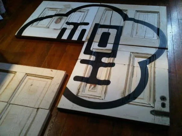 Large canvas with a black outline of a circular logo and vertical design elements, placed on a wooden floor near a set of painted cabinet doors.