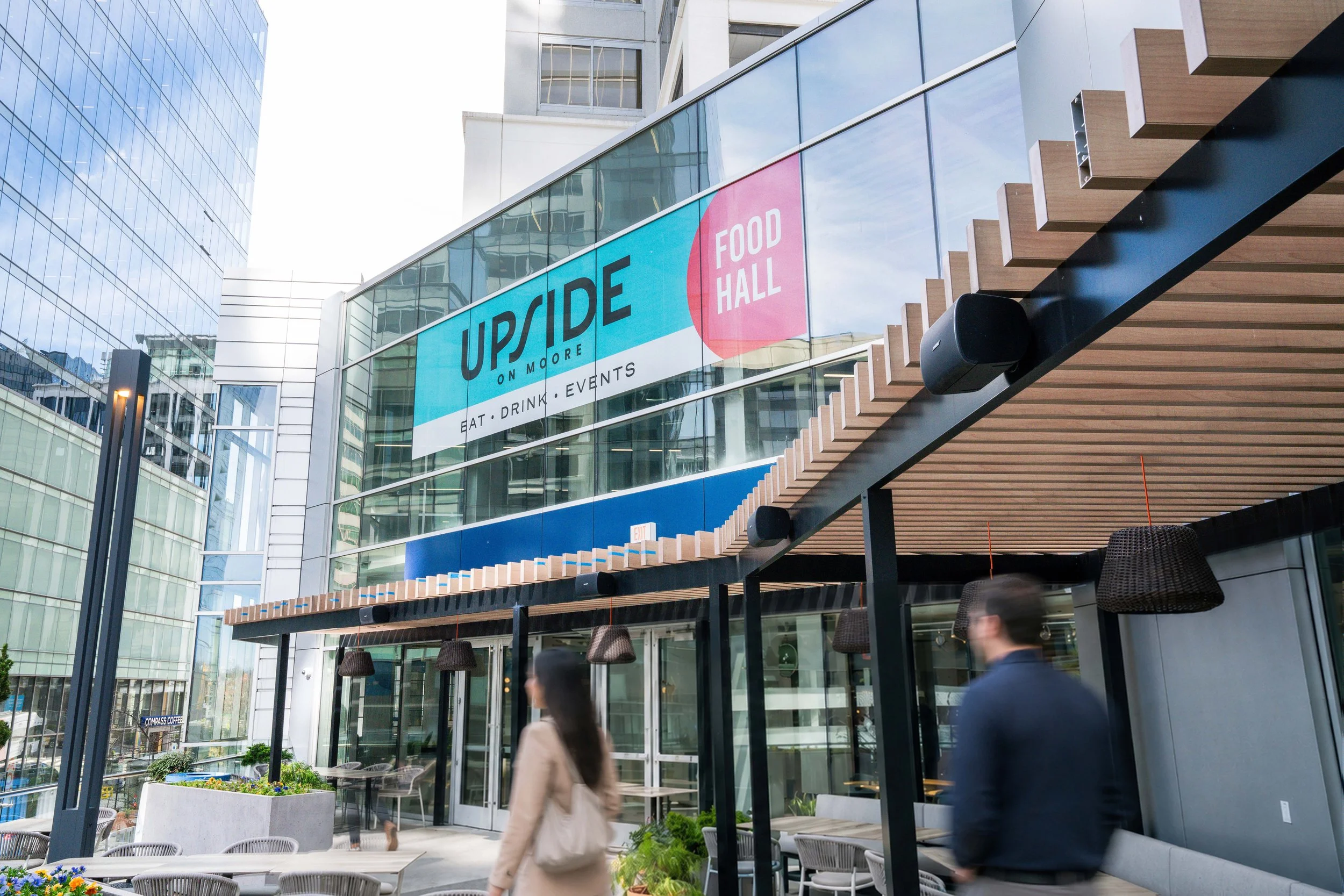 Modern glass building with a sign reading "Upside on Moore Food Hall". Outdoor seating area with tables, chairs, and potted flowers. Blurred people walking past.