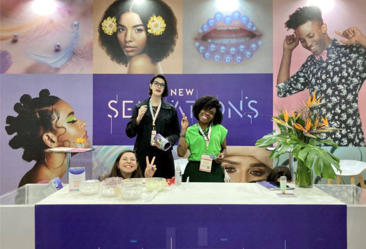 Three women are standing and sitting behind a white table at a beauty or cosmetics event, with a purple backdrop displaying large images of diverse women with different hairstyles and makeup. The women are smiling, and one is making a peace sign. The table has bowls of product samples, a flower arrangement, and promotional materials.