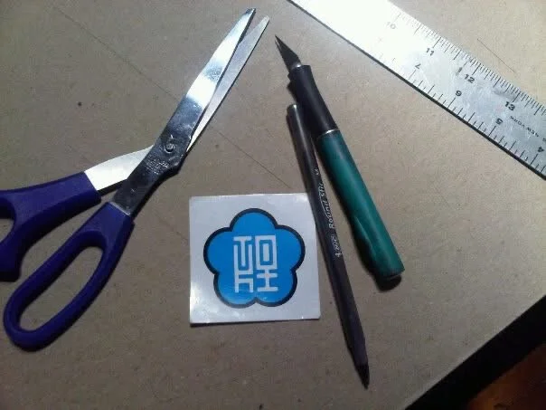 A pair of scissors, a pen, a mechanical pencil, a ruler, a sticker with a blue cloud and white text, all arranged on a beige surface.