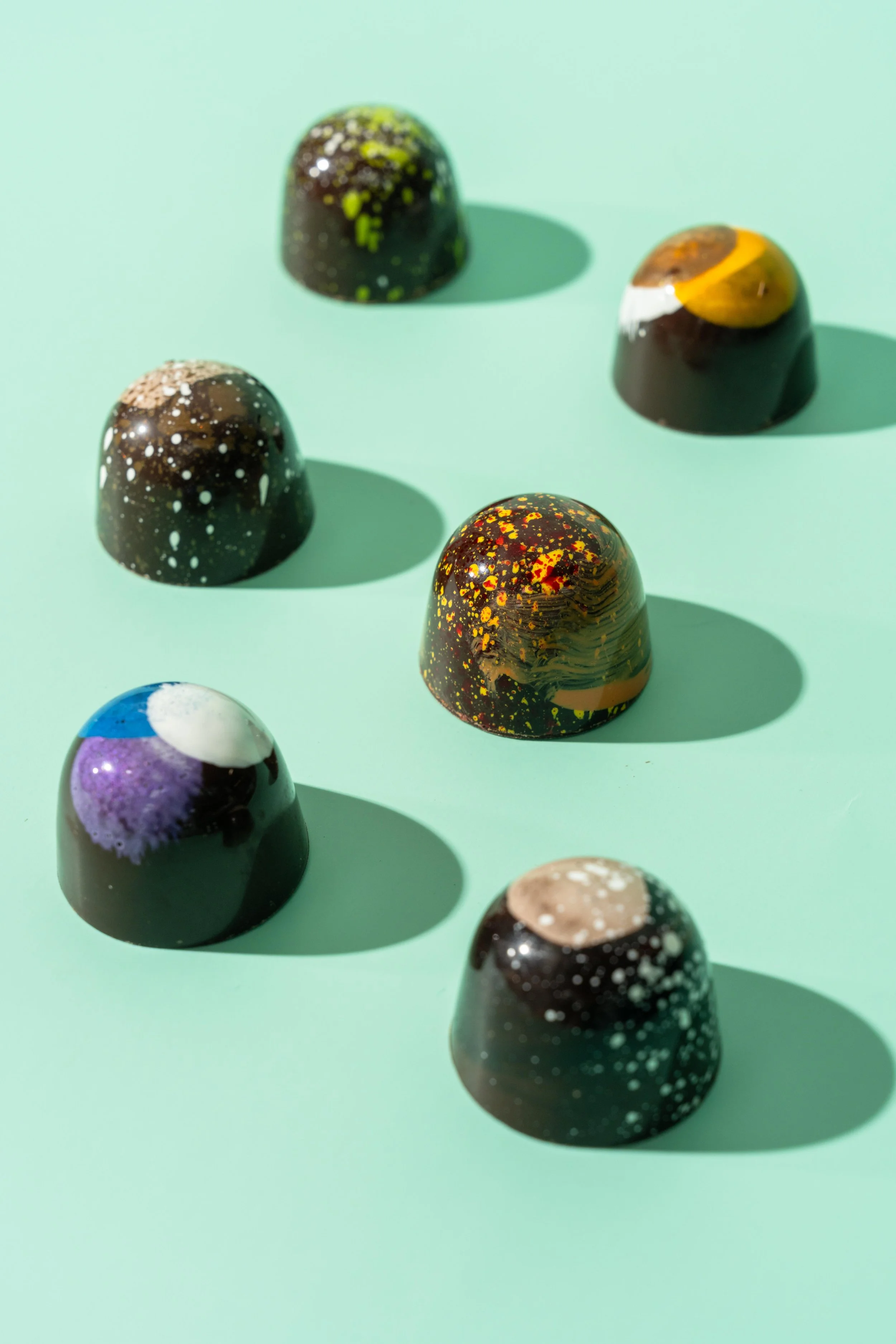 A group of colorful, speckled, dome-shaped chocolates arranged on a mint green surface, casting shadows.