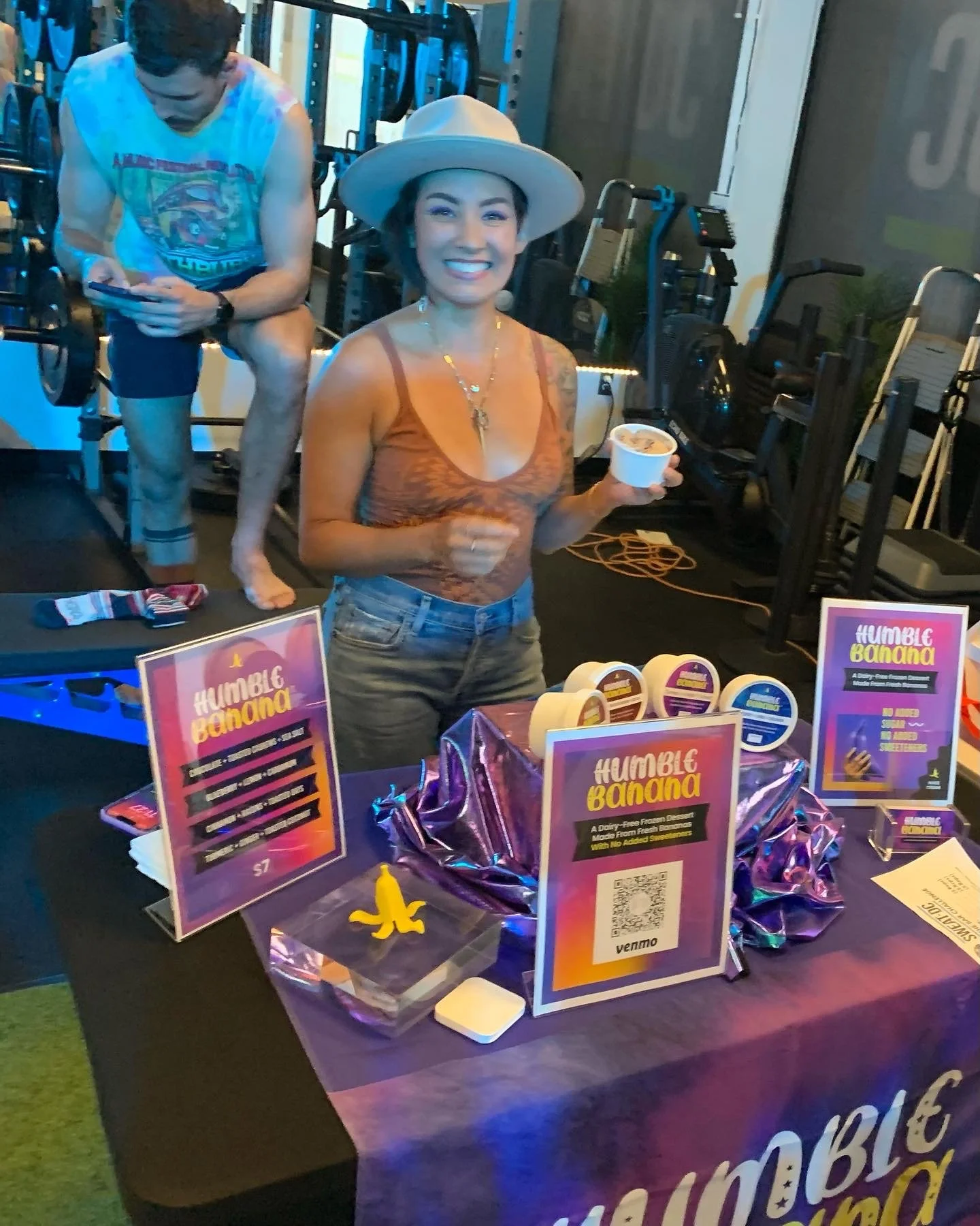 A smiling woman wearing a wide-brimmed hat and an orange top holding a bowl of frozen dessert at a booth promoting 'Humble Banana' with purple-themed packaging displays.
