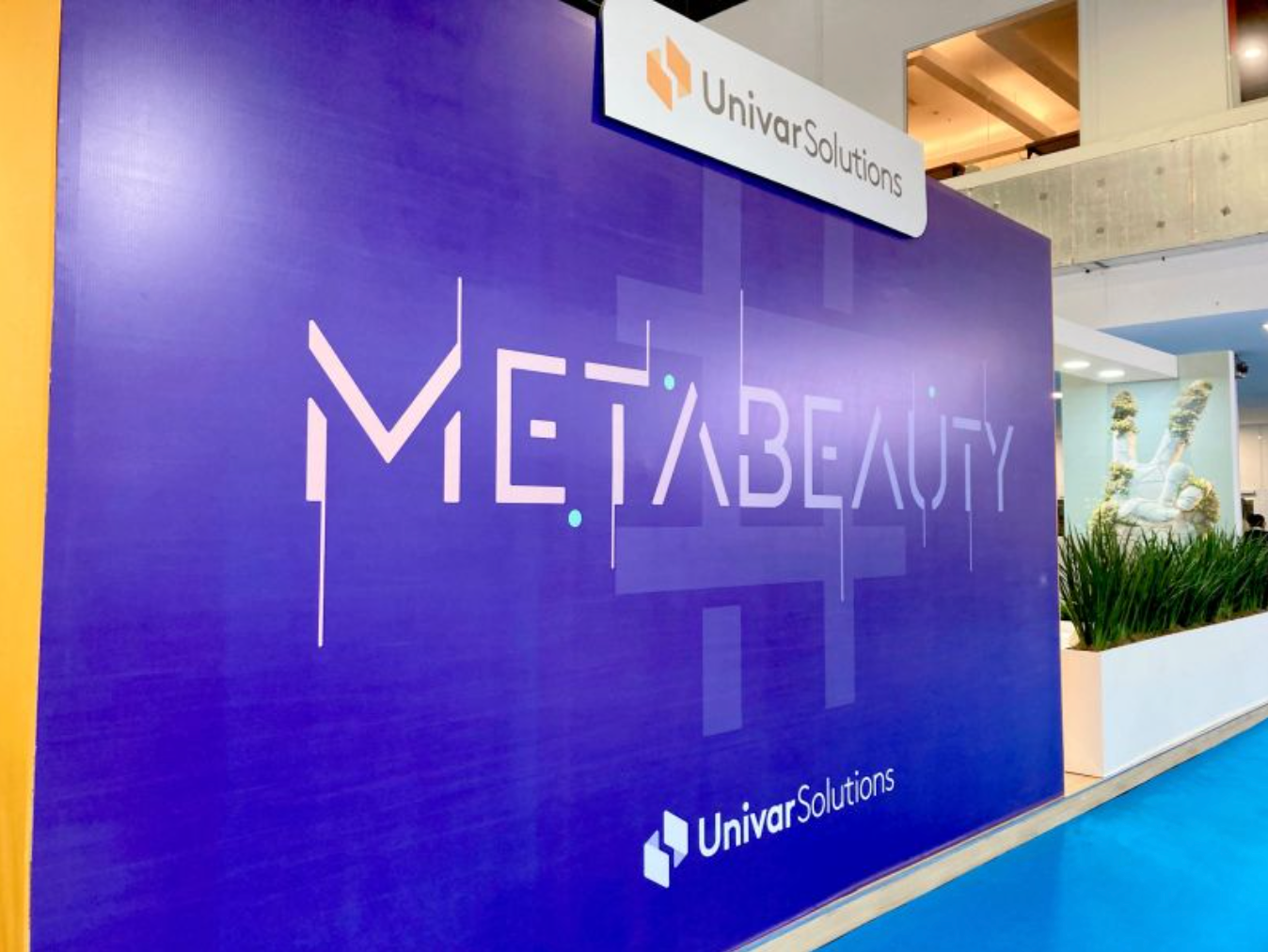 A large purple display board with the words 'Meta Beauty' in stylized text. The display is part of a booth or exhibition by Univar Solutions, whose logo appears at the top and bottom of the board. In the background, there are plants and a decorative chair.
