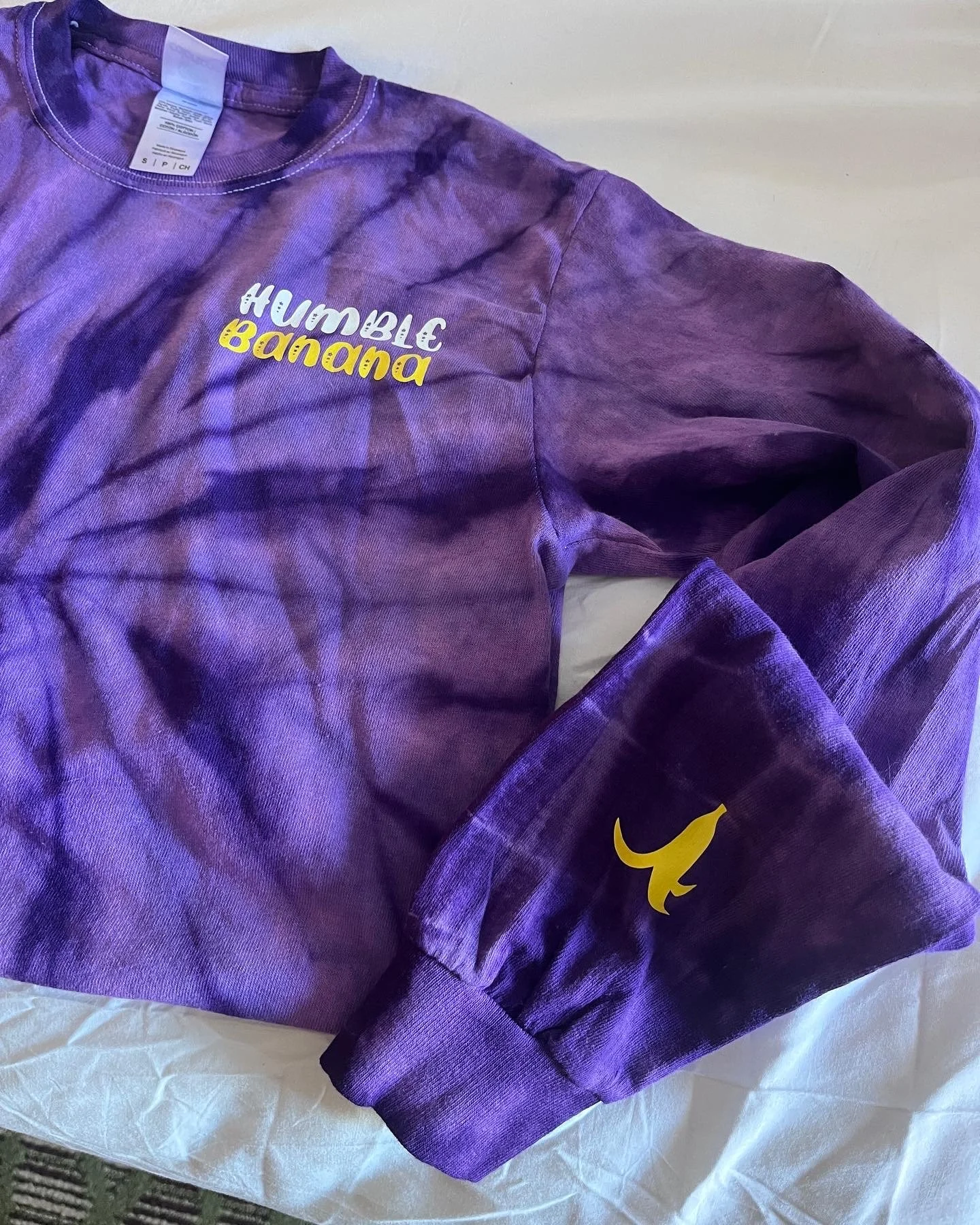 A purple tie-dye sweatshirt with white and yellow text that reads 'Humble Banana' and a yellow Nike swoosh logo on the sleeve.