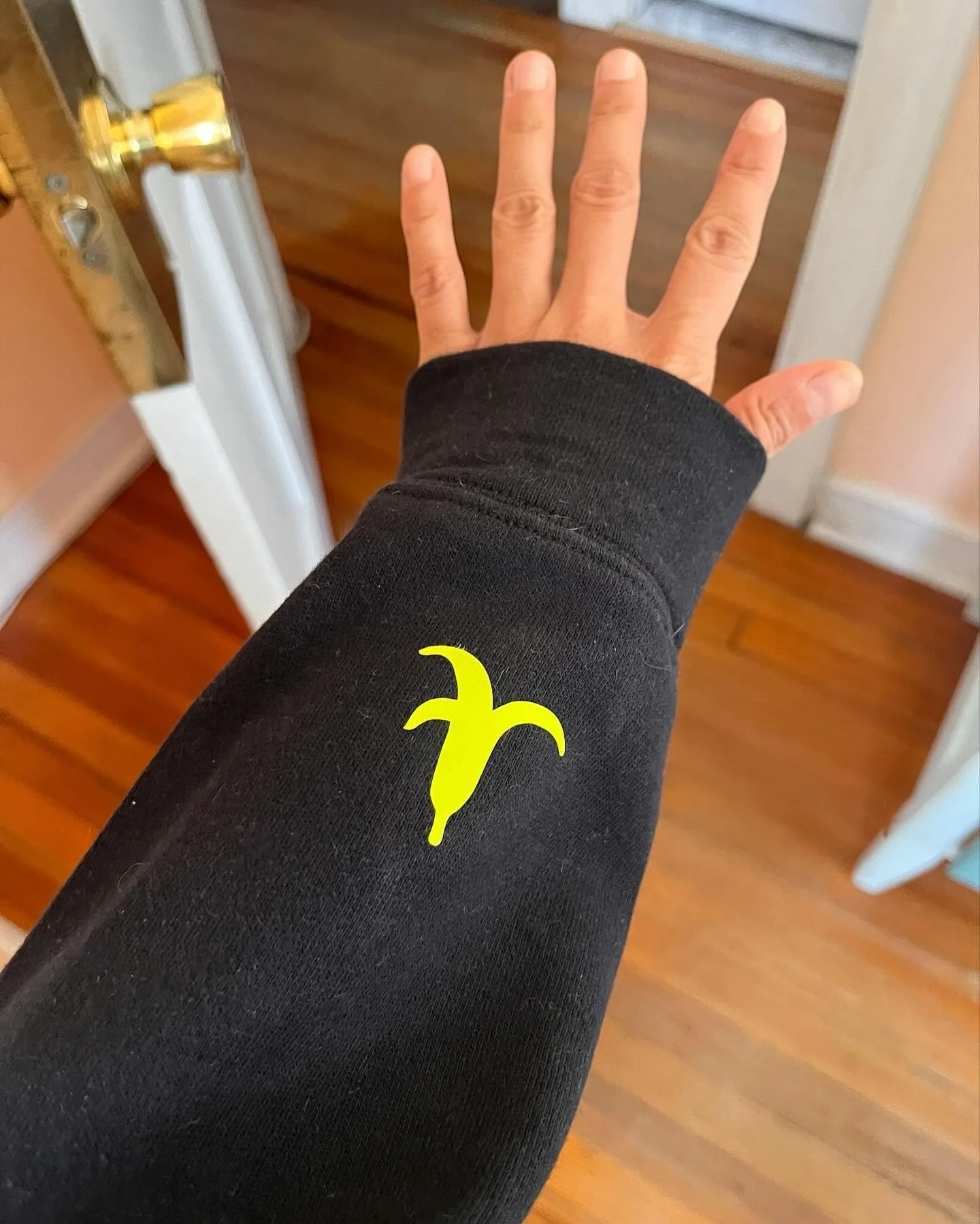 A person wearing a black long-sleeve shirt with a yellow logo on the wrist, extending their hand outward. The background shows a wooden floor, a white doorframe, and a brass-colored door handle.