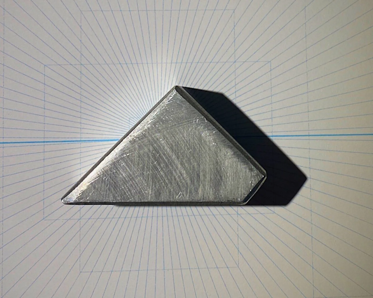 Close-up of a triangular-shaped metallic object with textured surface, placed on a sheet of graph paper with concentric radiating lines.