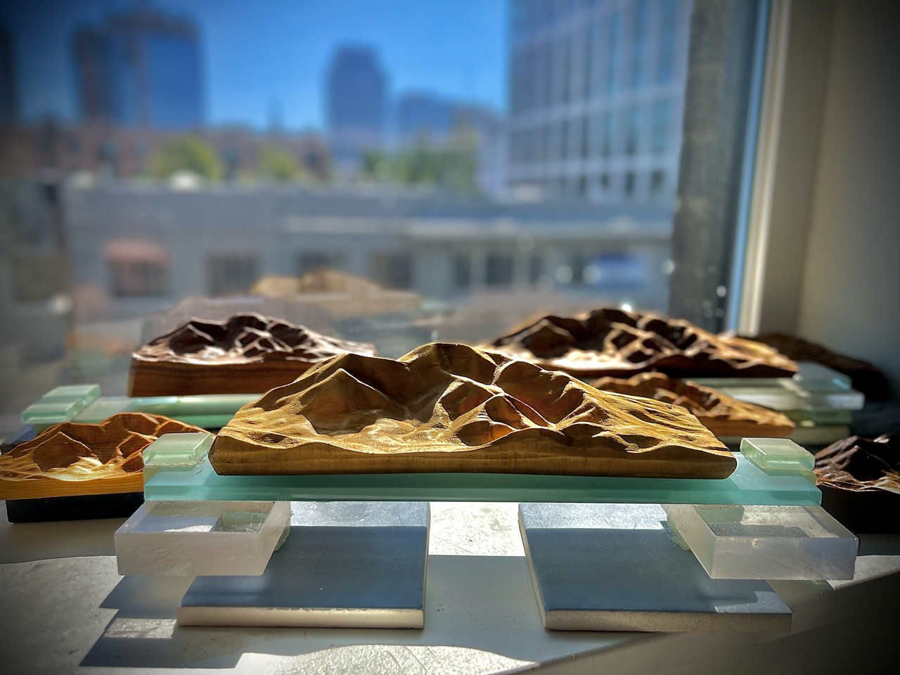 Multiple carved stone models of mountain landscapes arranged on glass and concrete display stands near a window with a city skyline in the background.