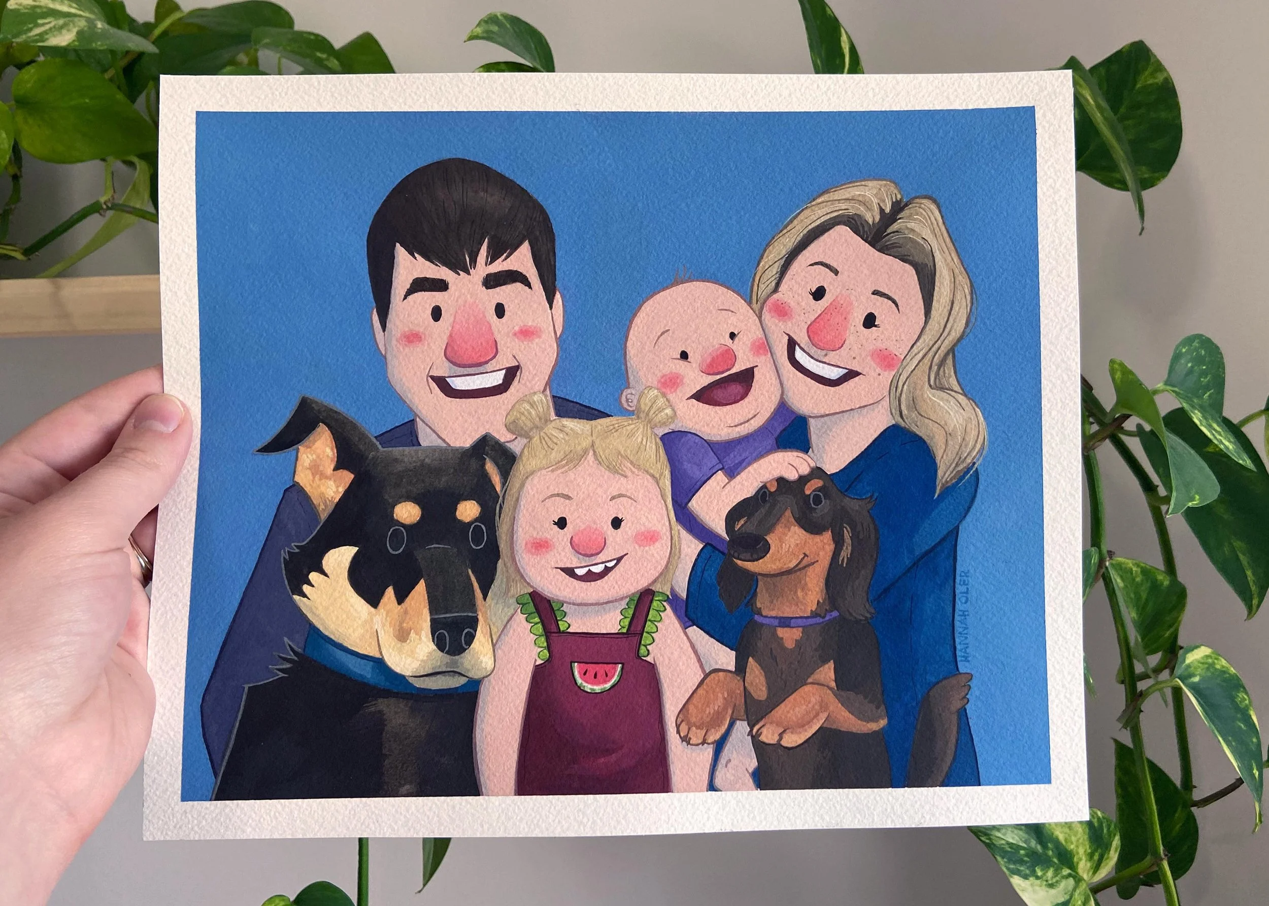 A hand holdes a painting of a family with two dogs, a mom and dad and two children, in front of a houseplant on a grey background.