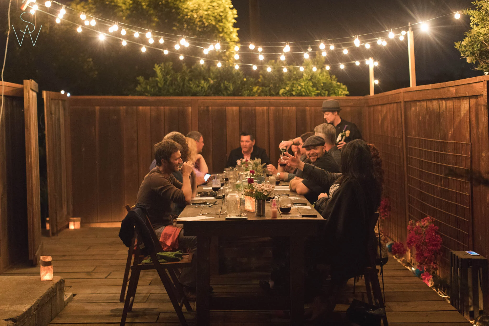 Group of people dining at a long outdoor wooden table during nighttime, illuminated by string lights, in a backyard with flowers and wooden fence.