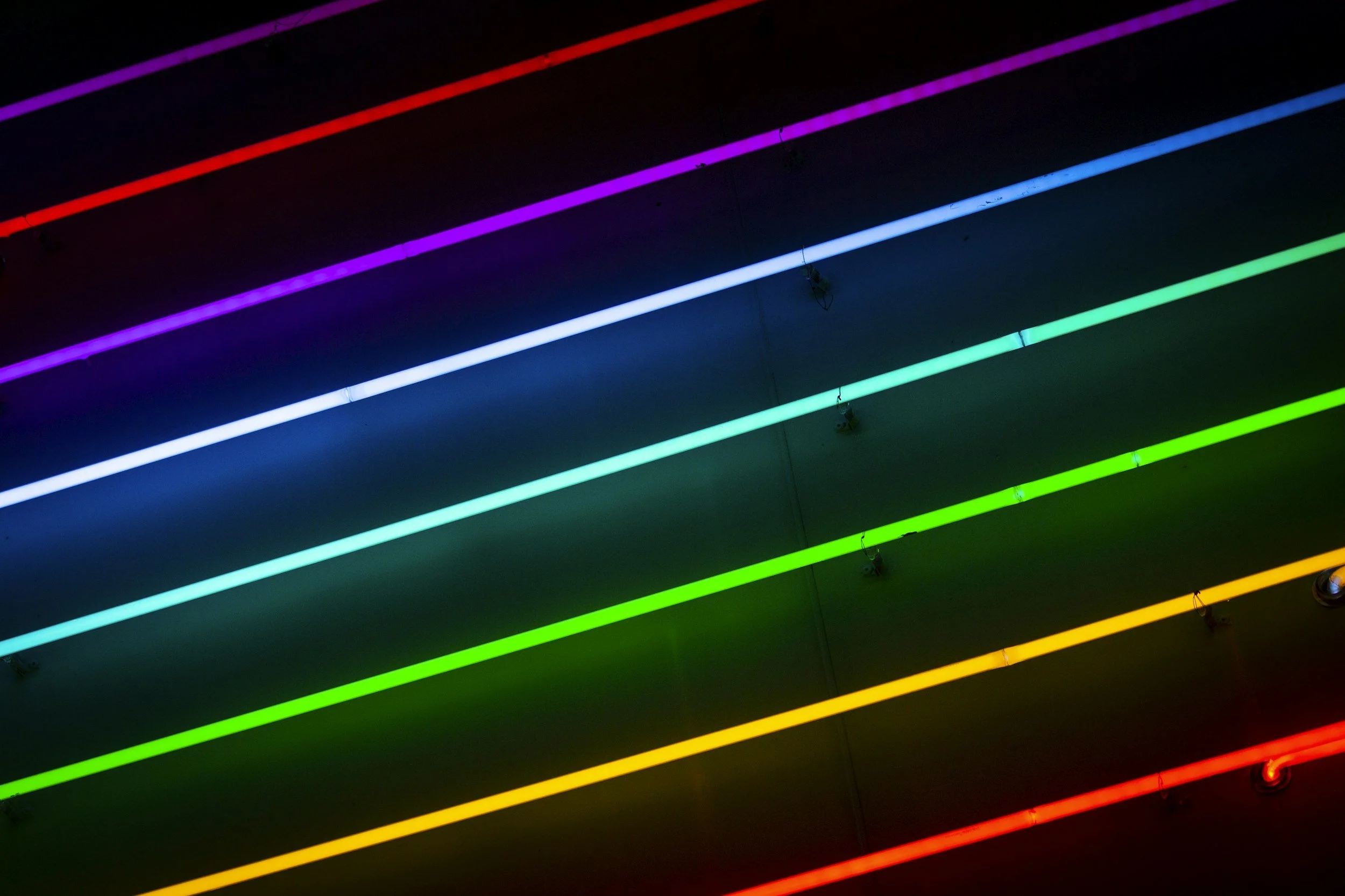 Multiple illuminated neon light strips in rainbow colors arranged diagonally against a dark ceiling.