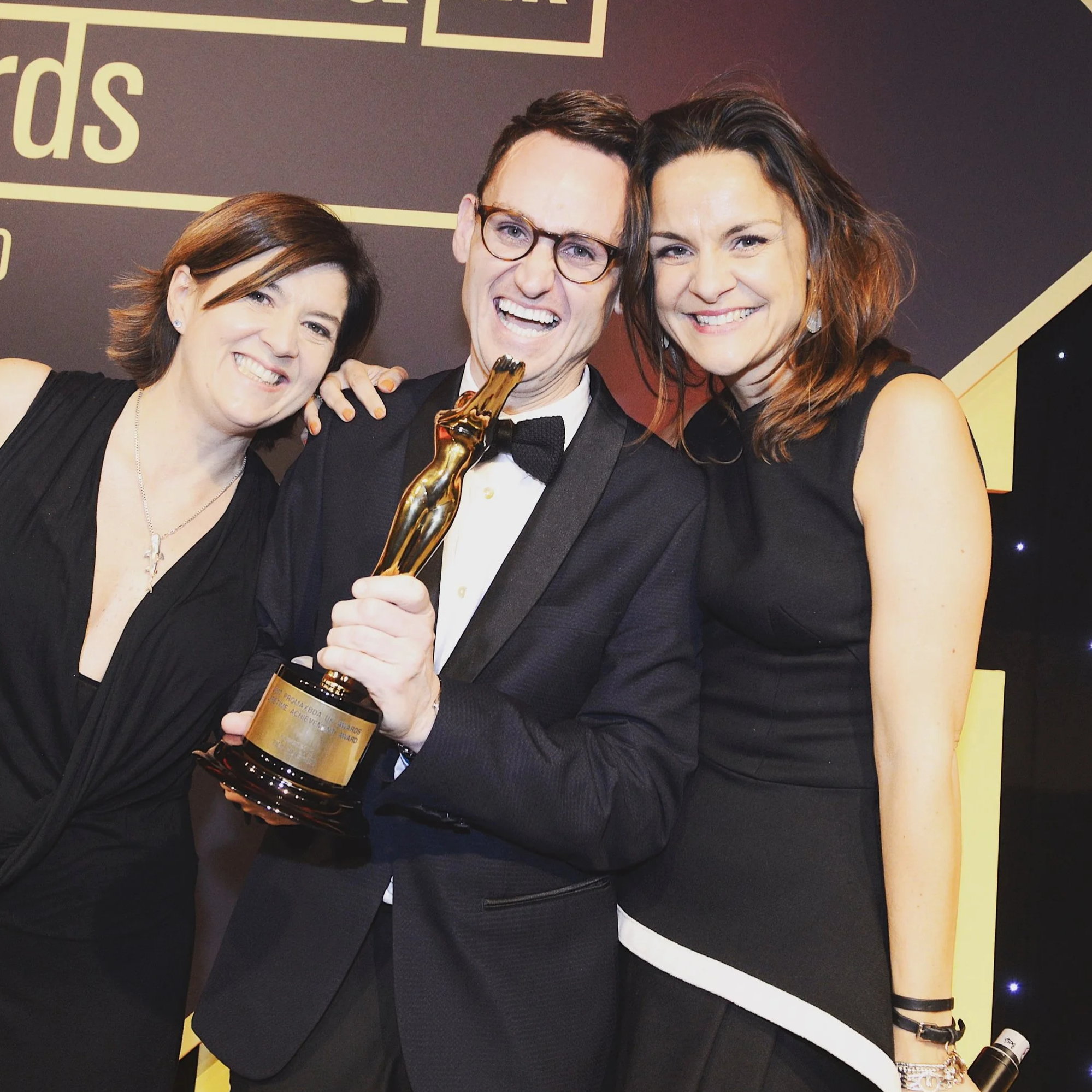 Three people celebrating an award at a formal event, with the man holding a gold trophy and smiling.
