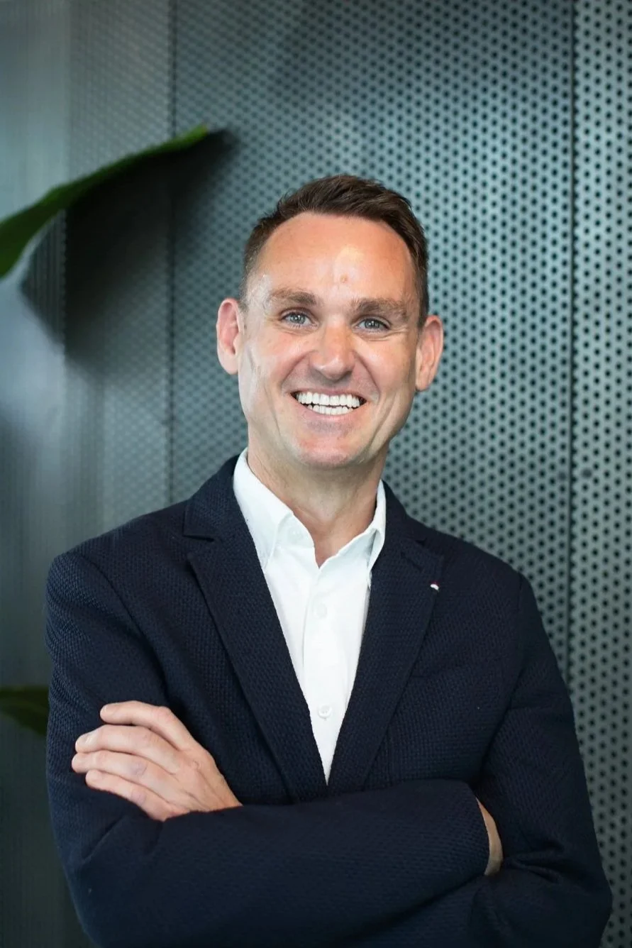 A smiling man in a business suit standing with arms crossed in front of a modern, perforated metal wall.