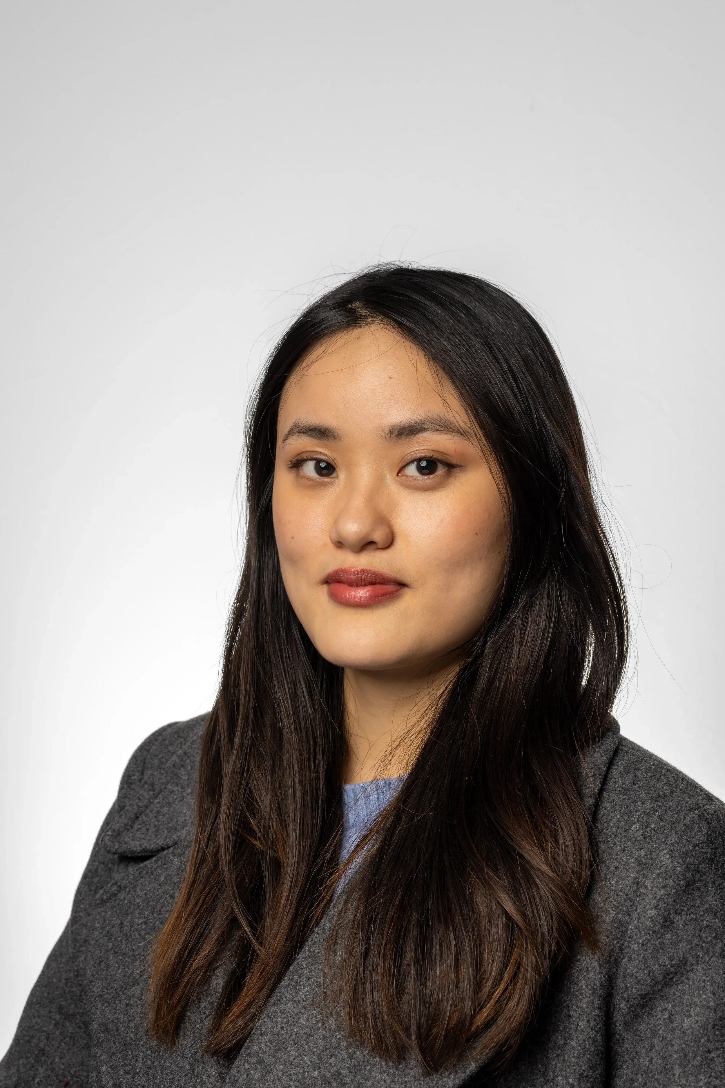A young woman with long dark hair, wearing a gray coat and a light blue top, looking at the camera against a plain light gray background.