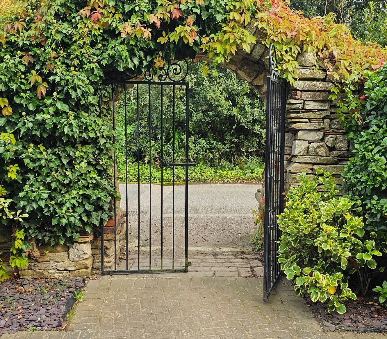 A black wrought iron gate attached to a stone archway covered in green and reddish foliage, leading to a paved street with greenery in the background.