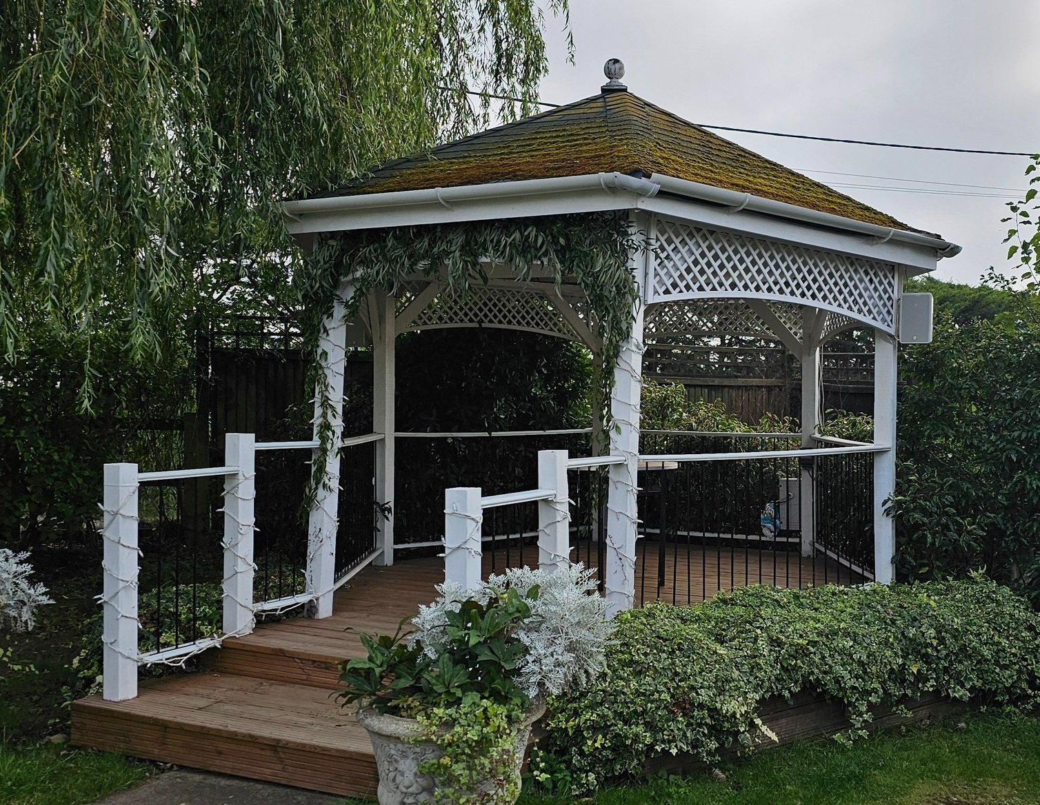 White gazebo with a brown wooden floor and black metal railing, surrounded by green bushes and trees, with an overcast sky in the background.