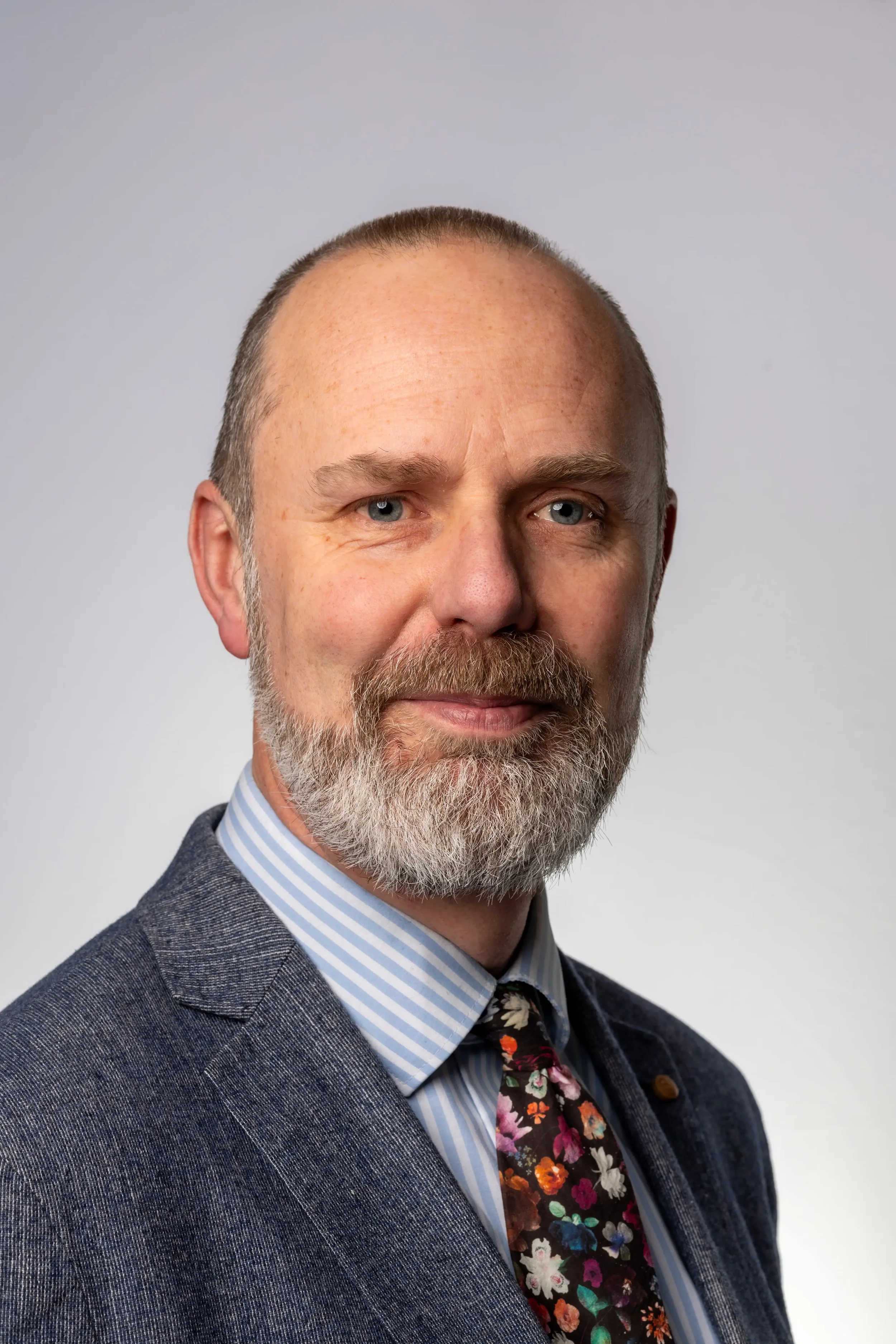 A middle-aged man with a beard and short hair, wearing a suit with a striped shirt and a floral tie, posing against a plain background.