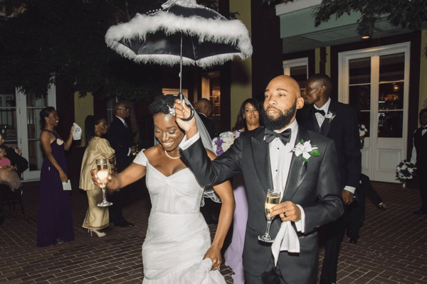A joyful couple at a wedding, holding a feathered umbrella while celebrating with guests.