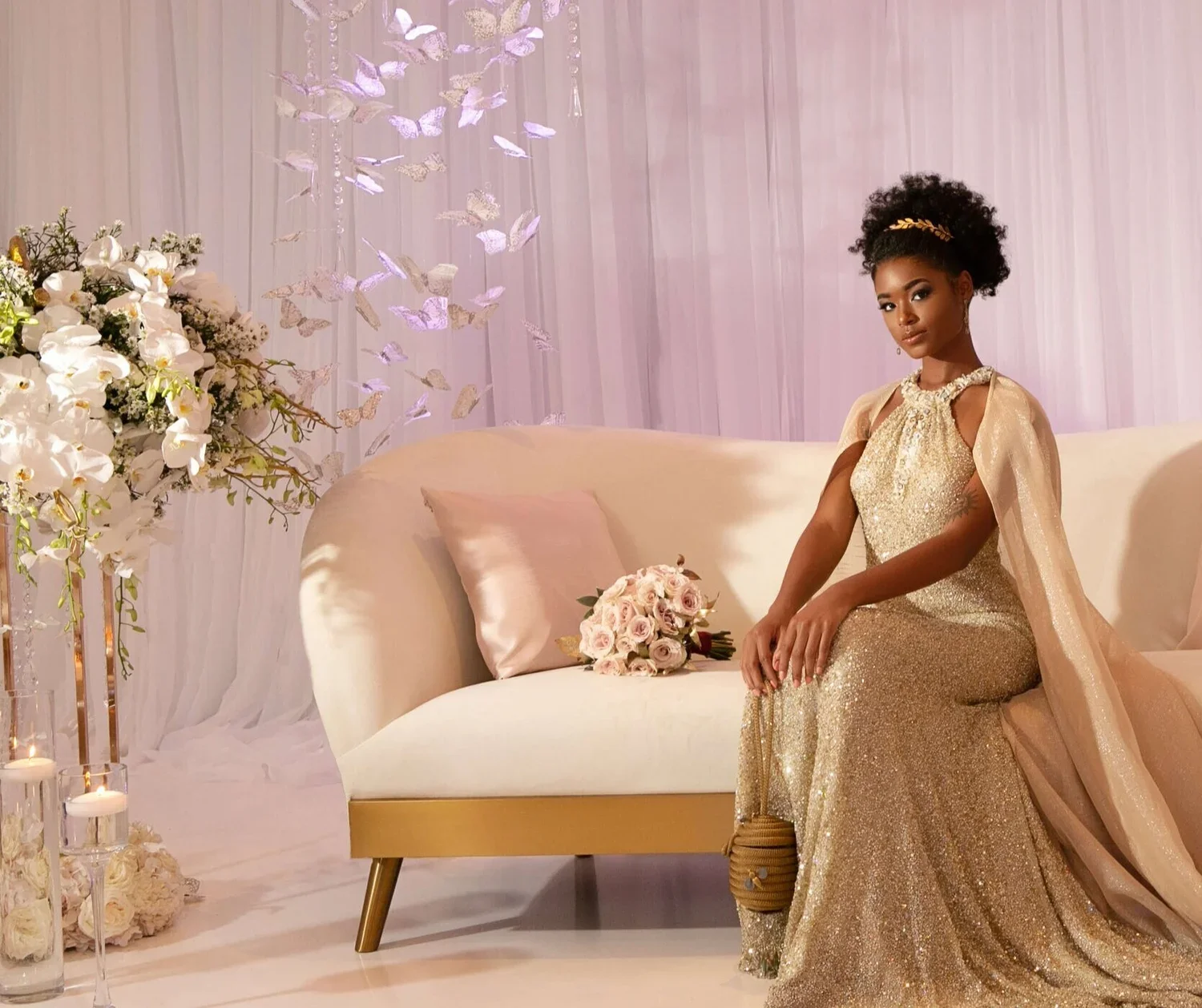 Bride wearing a shimmering gold gown with a matching cape, seated gracefully on a white and gold sofa surrounded by white floral arrangements, candles, and a soft pink bouquet in a romantic setting.