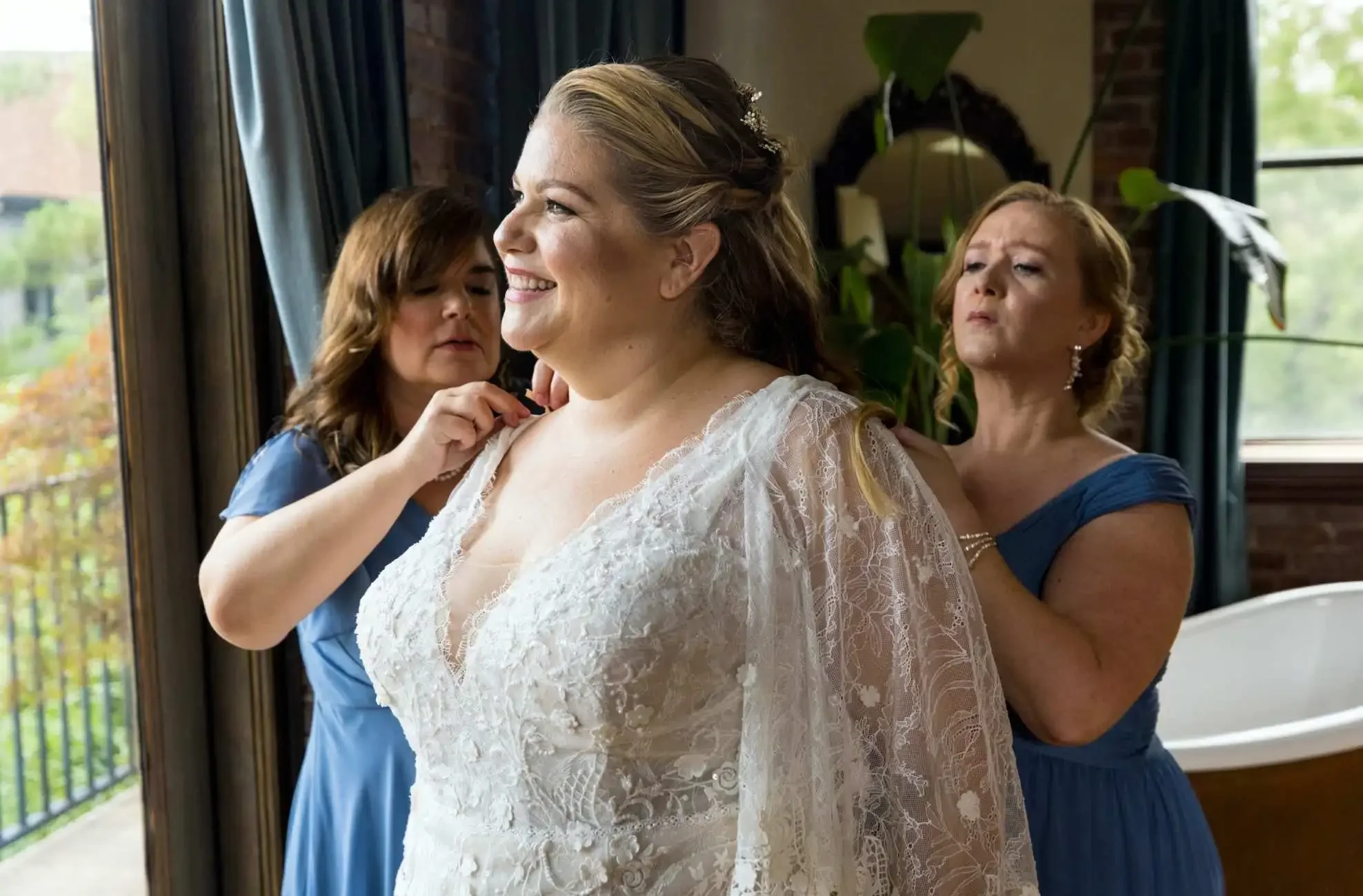 Bride in a lace gown smiles near a window as two bridesmaids in blue dresses help fasten her dress before the ceremony.