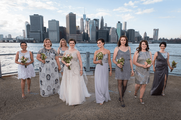 Eight women wearing colorful dresses pose together by the waterfront with a skyline backdrop.