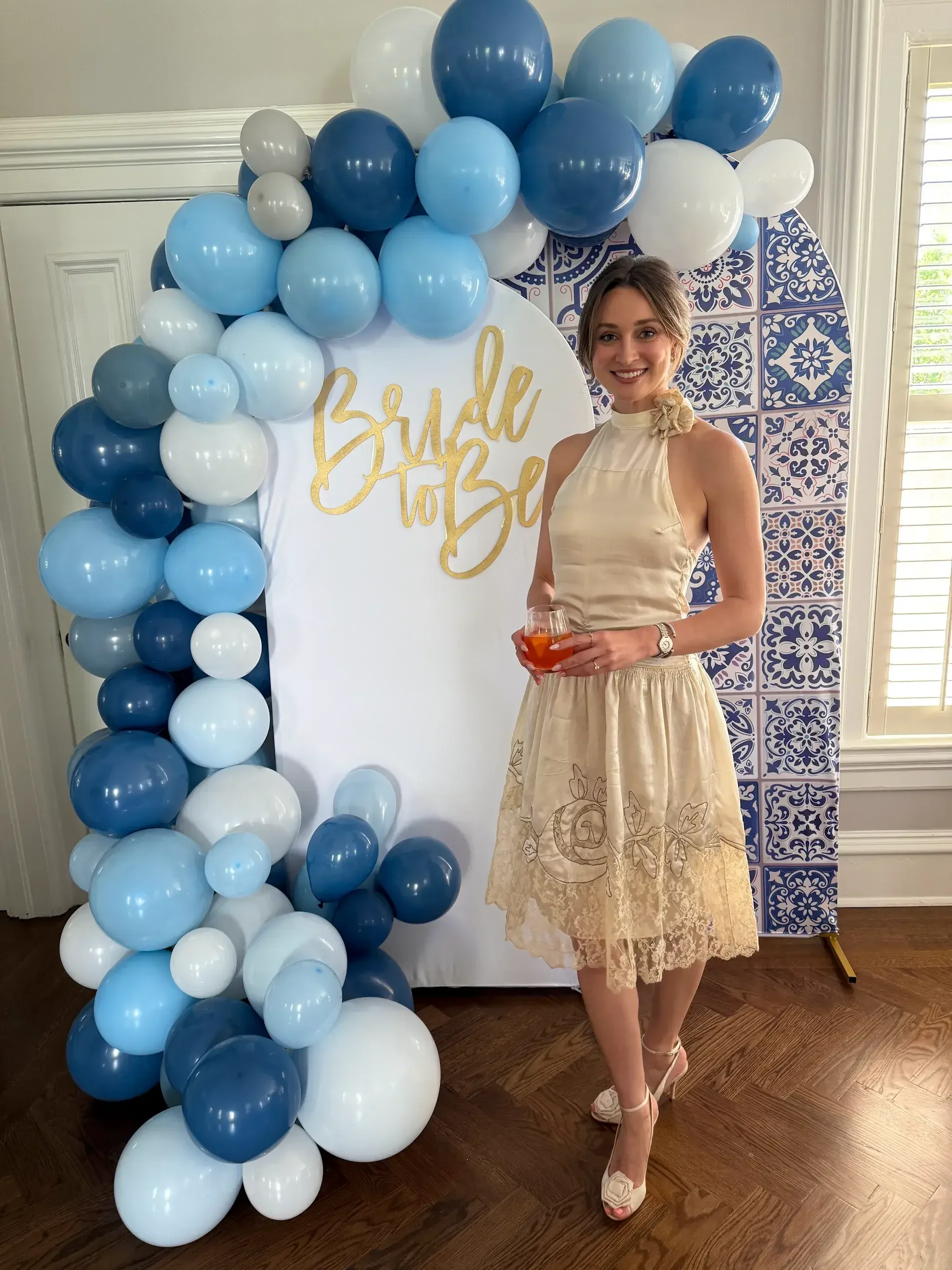 Bride-to-be standing beside a blue and white balloon arch with gold “Bride to Be” sign, wearing a beige lace dress and holding a drink at her bridal shower.