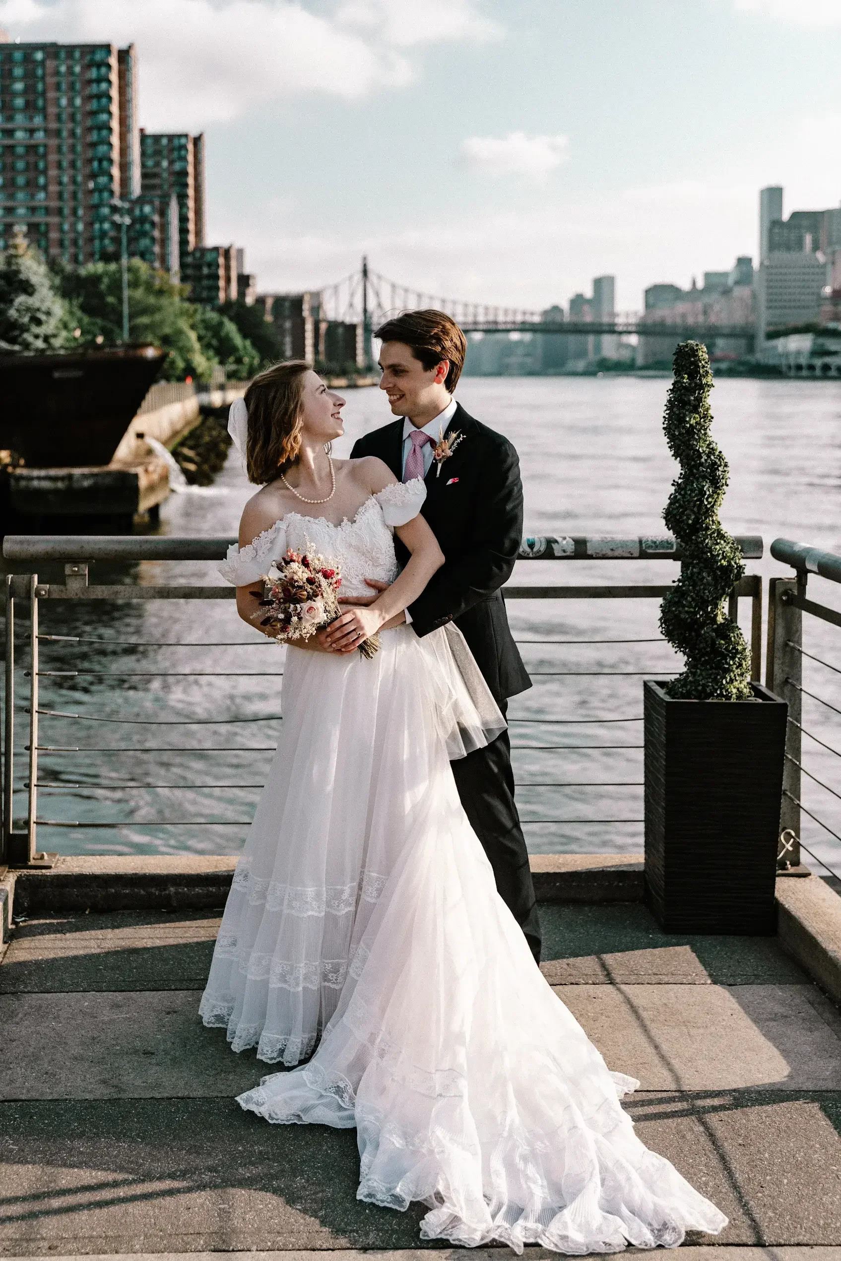 ride in an off-the-shoulder lace gown embraces her groom by the waterfront, wearing a beautifully tailored dress that flows elegantly in the evening light.