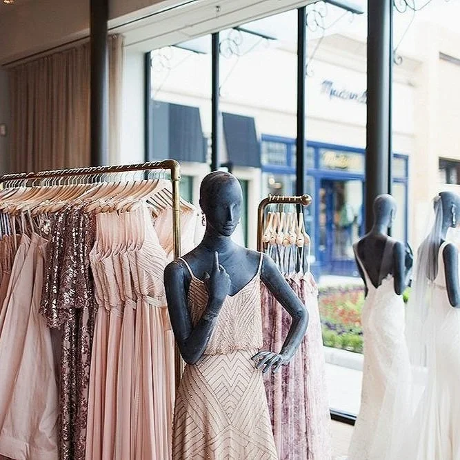Elegant bridal boutique interior featuring mannequins dressed in blush and champagne gowns, with racks of sequin and chiffon bridesmaid dresses displayed near large windows.