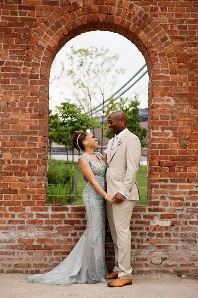 Bride in a light blue embroidered gown and groom in a beige suit standing hand in hand under a rustic brick archway, smiling at each other with greenery and a bridge in the background.
