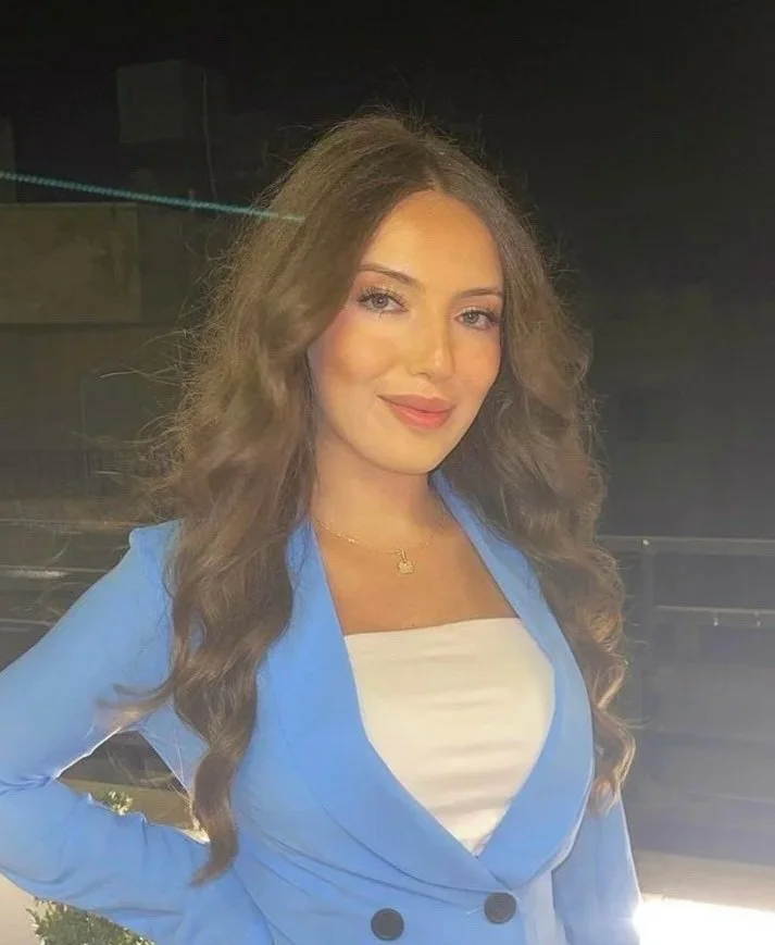 A young woman with long, curly brown hair, wearing a light blue blazer over a white top, and a small gold necklace. She is smiling softly and has a confident pose with one hand on her hip.