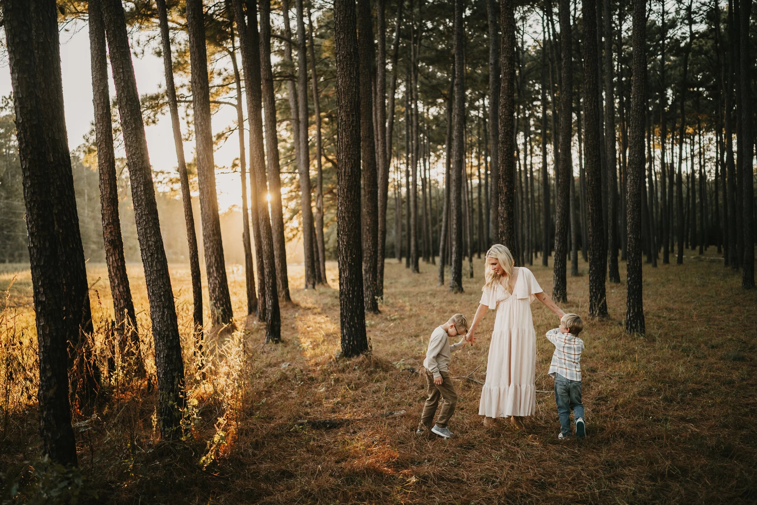 A woman and two children holding hands and playing in a sunlit forest with tall trees and a grassy floor during sunset.