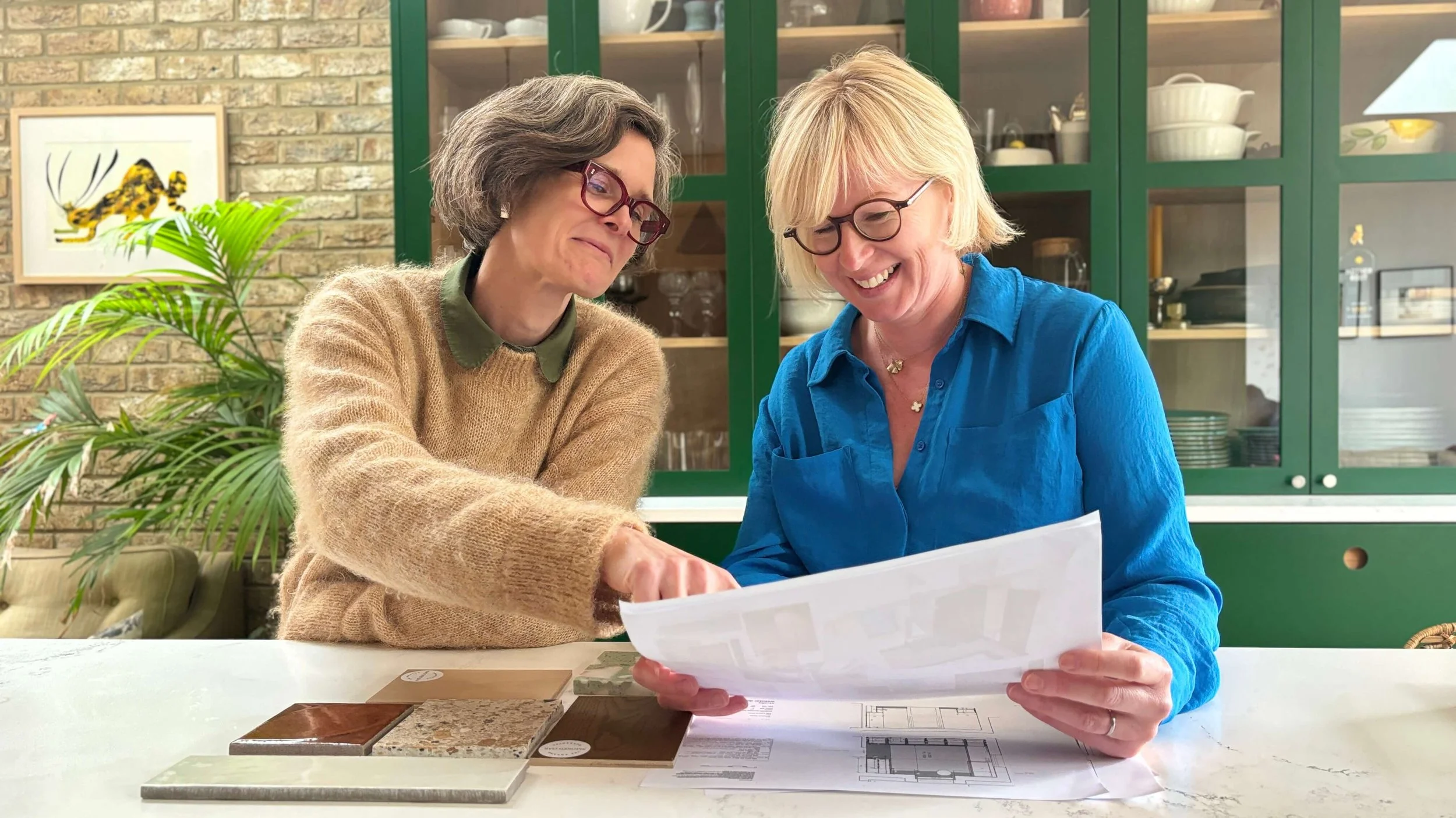 Two women looking at a blueprint or architectural plan together in a kitchen or dining area with green cabinets and brick wall in the background.