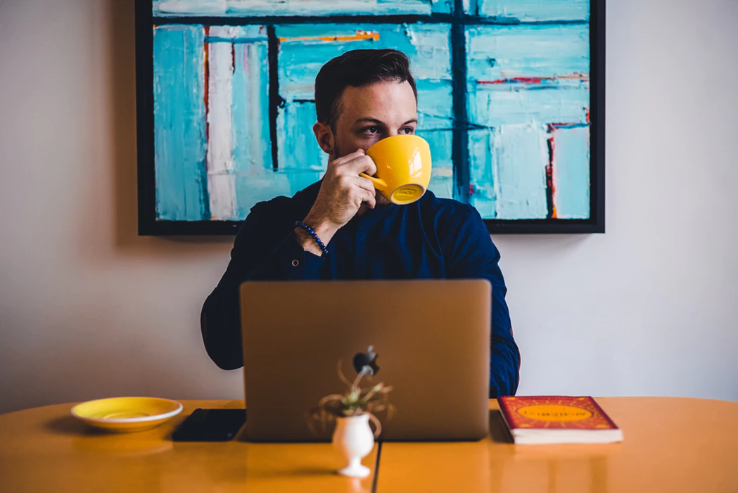 man drinking from a mug while doing remote work with a laptop