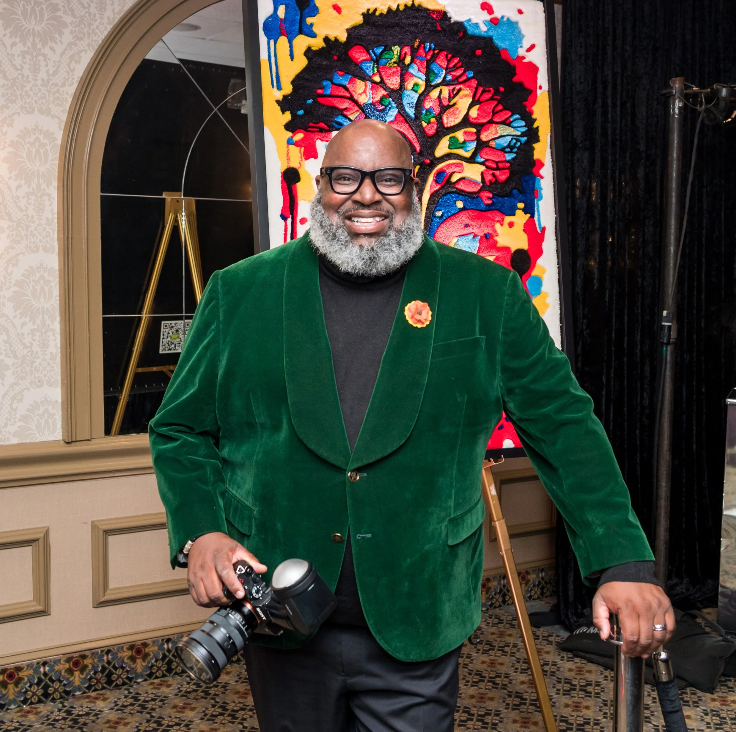 A man with glasses and a gray beard smiling, holding a professional camera, standing in front of a colorful abstract painting. He is wearing a green velvet blazer with a flower pin, black shirt and pants.