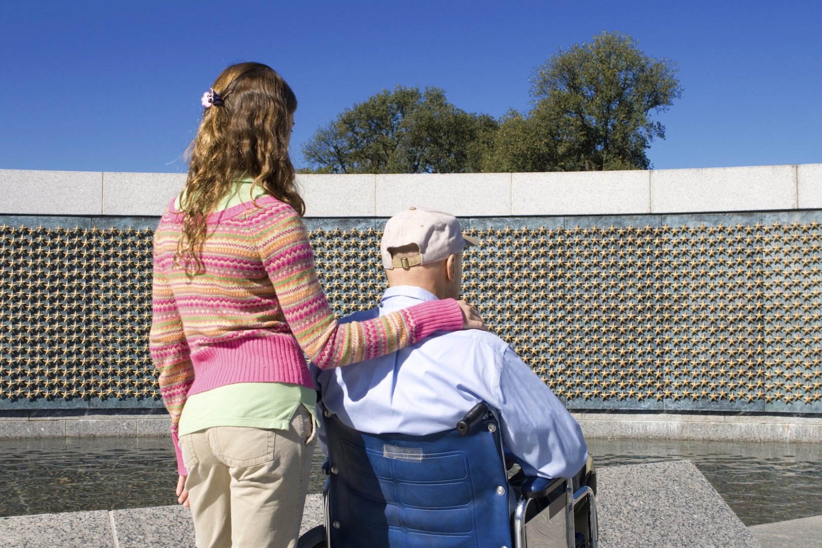  A girl with her arm around an elderly man in a wheelchair. They both look at a veterans memorial with hundreds of stars on it. 