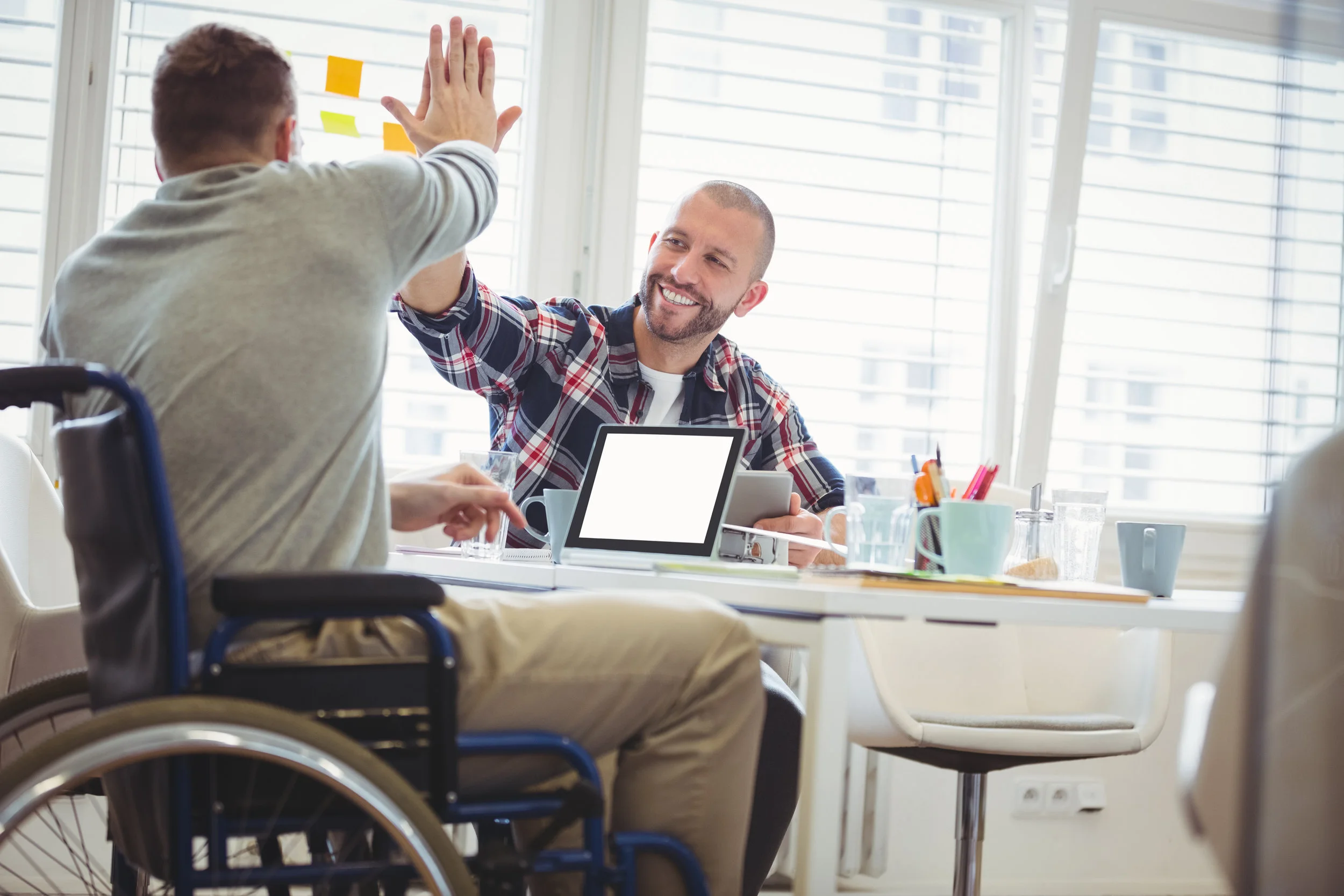 Image: A man using a wheelchair high-fives another man who is seated at a table with him. The man using the wheelchair is typing on a laptop. (Source: https://shutr.bz/2DxKM0i)