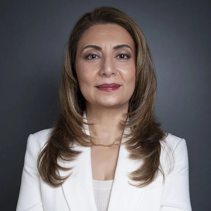 A woman with shoulder-length brown hair, wearing a white blazer and a gold necklace, standing against a dark background.