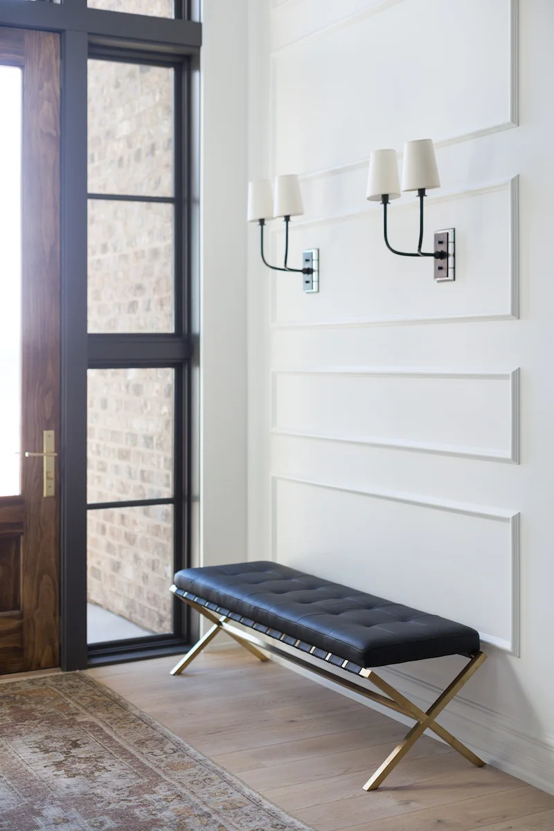A welcoming front entry with bench, wall sconces and wall panel details from a Rural Alberta Country House, Interior Design by Calgary Design Studio Nyla Free Designs Inc, Architecture: Dejong Design Associates, Photography: Phil Crozier, #nylafreedesigns #calgaryinteriordesigners #blackdoors #wallpanelling #countryhouse #styledpretty #frontentryenvy