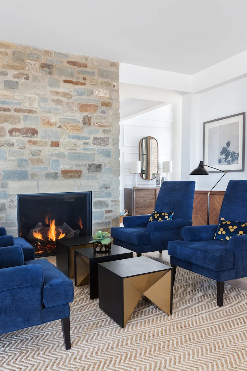 A sitting room with 4 navy chairs in front of a stone fireplace from a Rural Alberta Country House, Interior Design by Calgary Design Studio Nyla Free Designs Inc, Architecture: Dejong Design Associates, Photography: Phil Crozier #calgaryinteriordesign #calgaryinteriordesigner #nylafreedesigns #sittingroominspiration #countryhouse