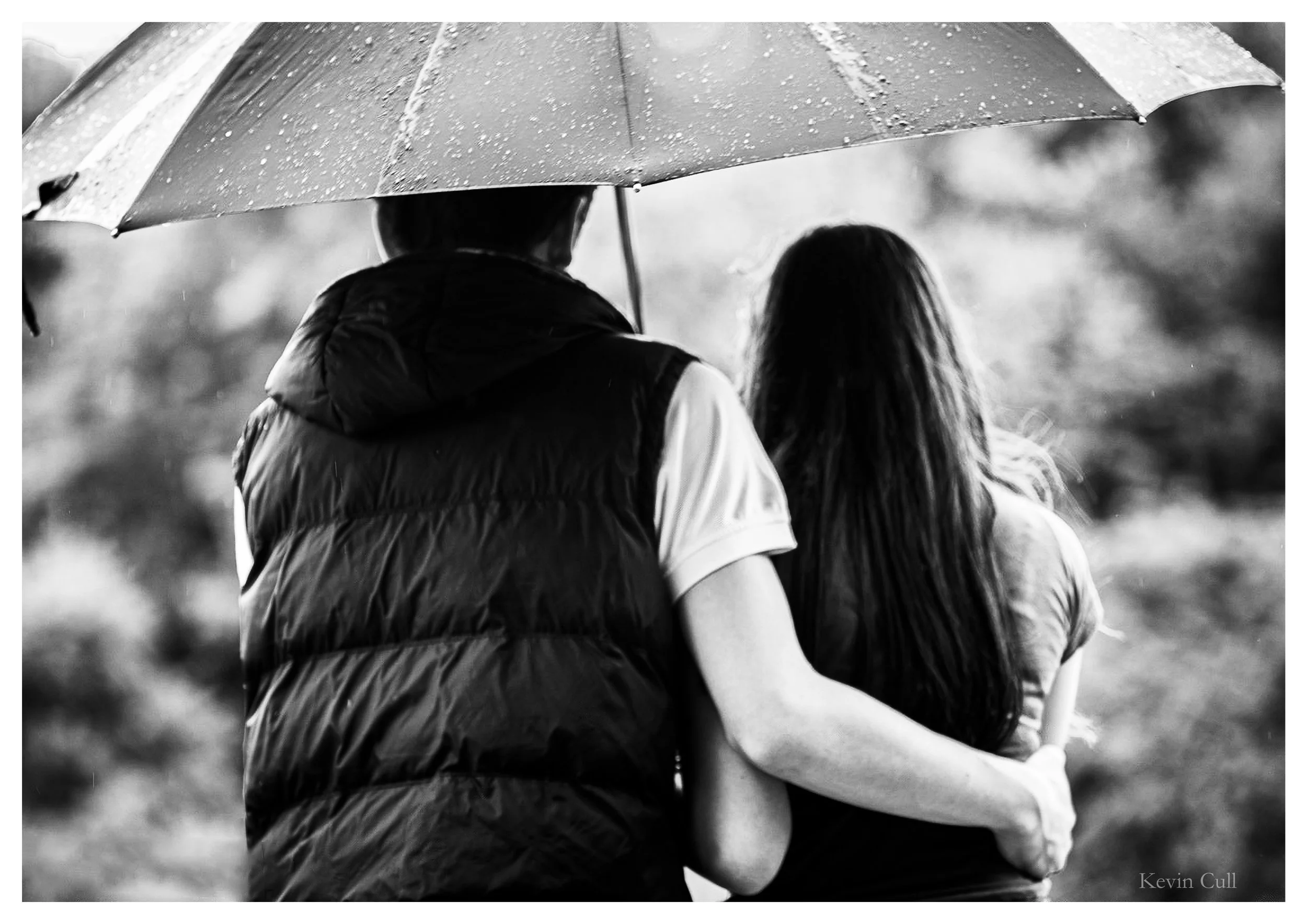 A black and white photo of a couple walking in the rain under an umbrella. The man is wearing a gillet and the woman has long hair. They are walking closely together with their backs to the camera.