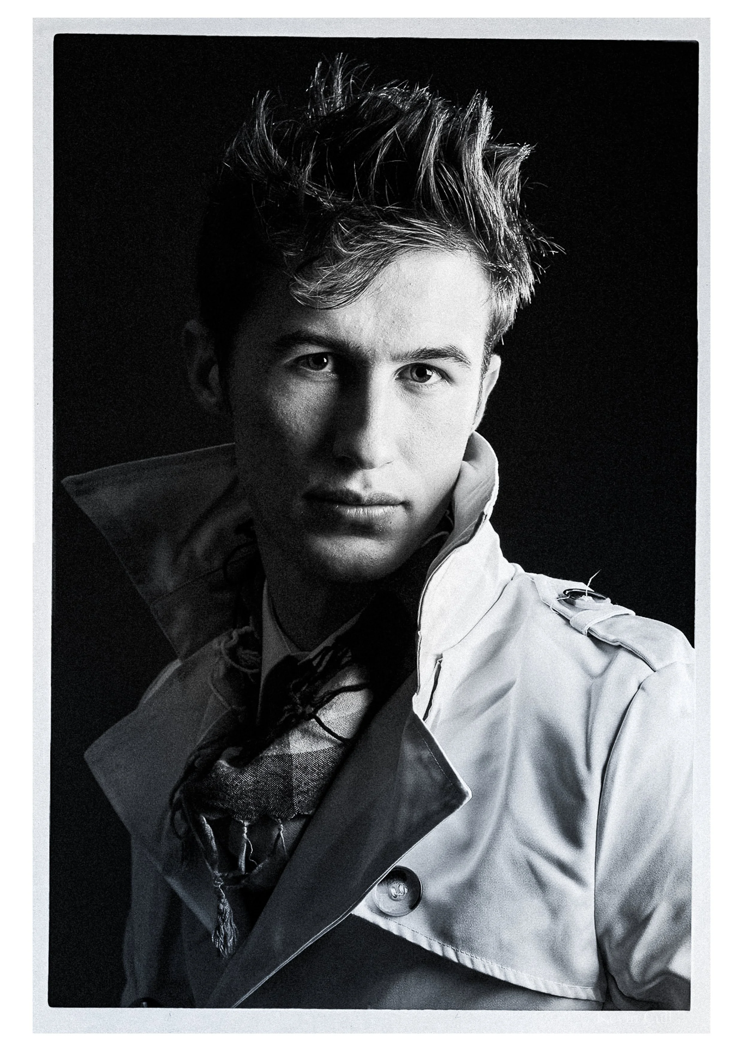 Black and white portrait of a young man with styled hair, wearing a jacket and a patterned scarf underneath, set against a dark background.
