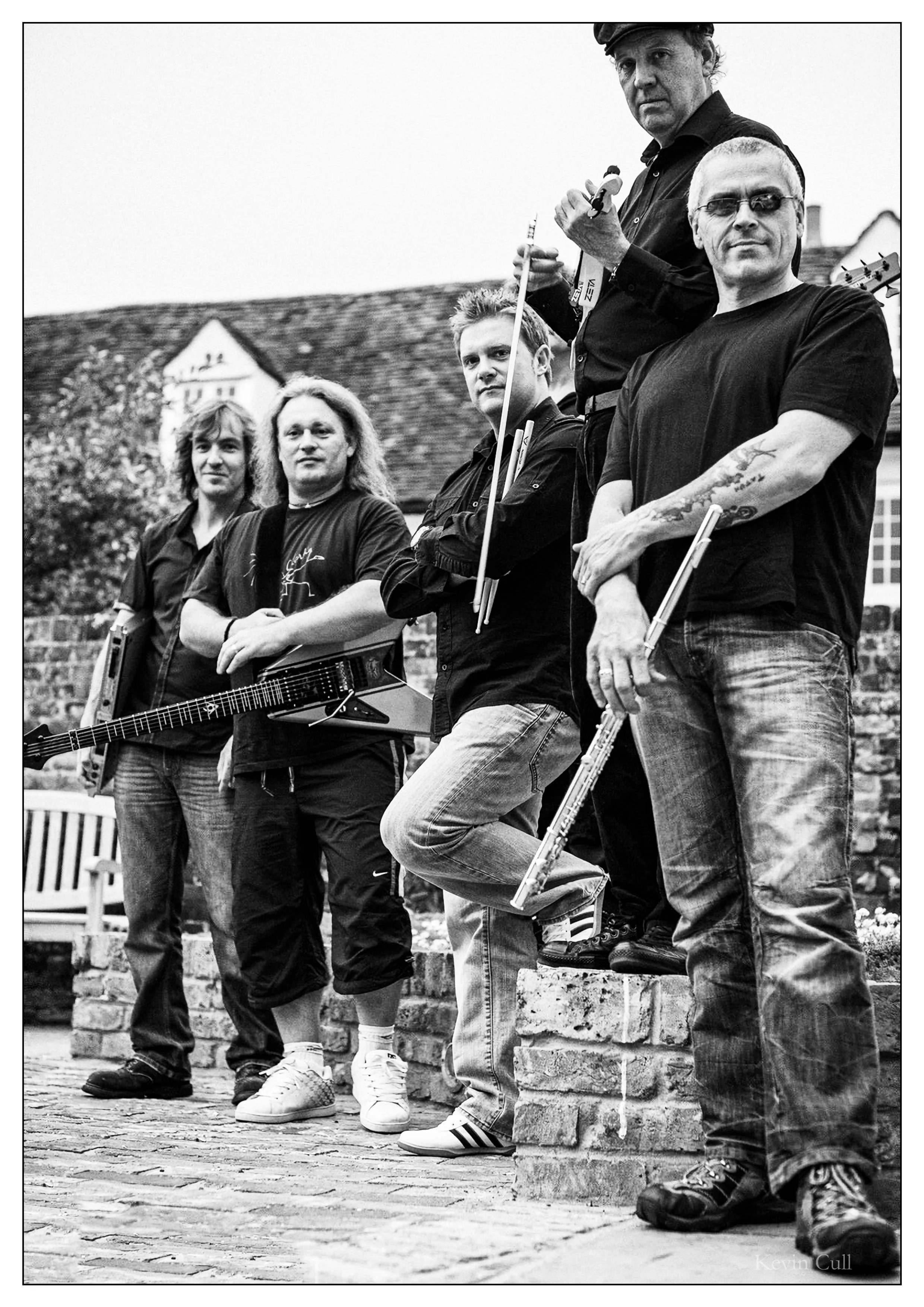 Black and white photo of five men standing outdoors against a brick wall and a house with a pitched roof. The men are holding musical instruments, including a guitar, a clarinet, and a drumstick. They are dressed casually, with some wearing t-shirts 