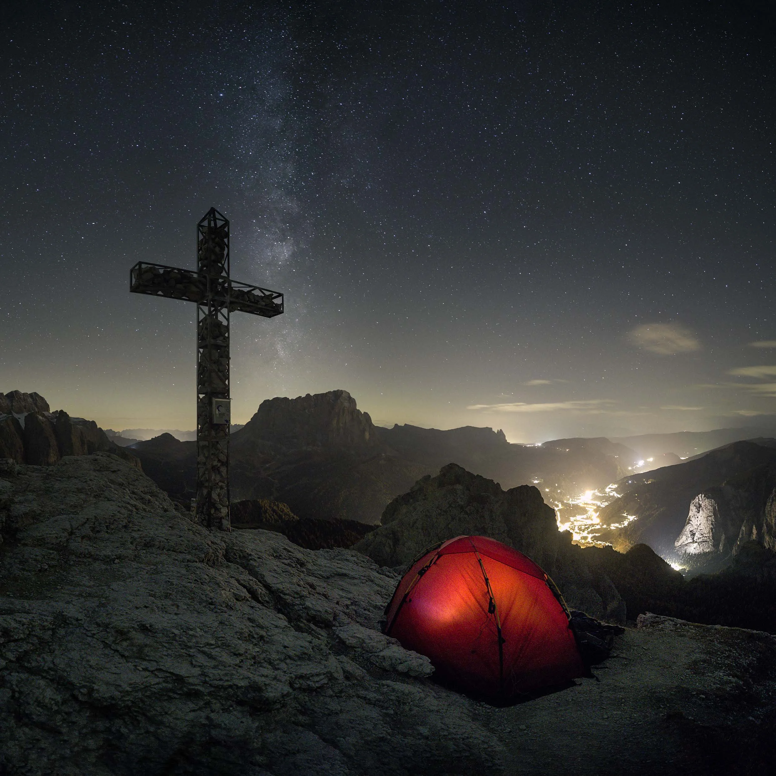 Hiker camping at the top of Gran Cir next to the mountain cross under the night sky showing the milky way galaxy. 
