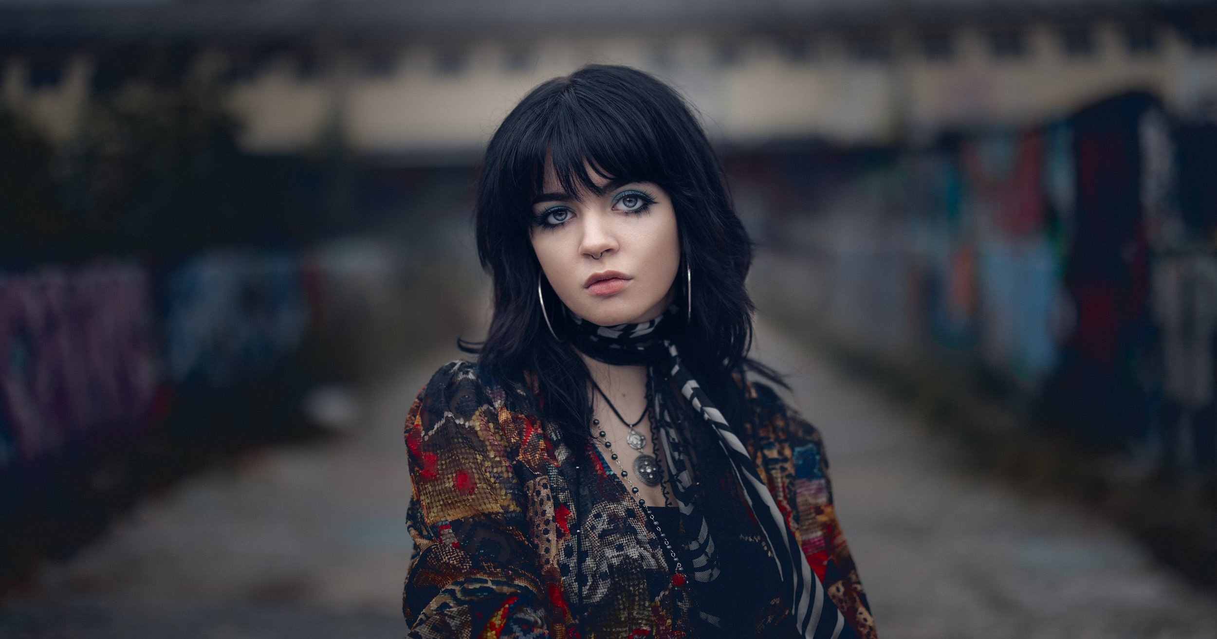 Young, beautiful girl with large, sad eyes, wearing a colorful, alternative-style shirt in front of graffiti walls in Munich. 