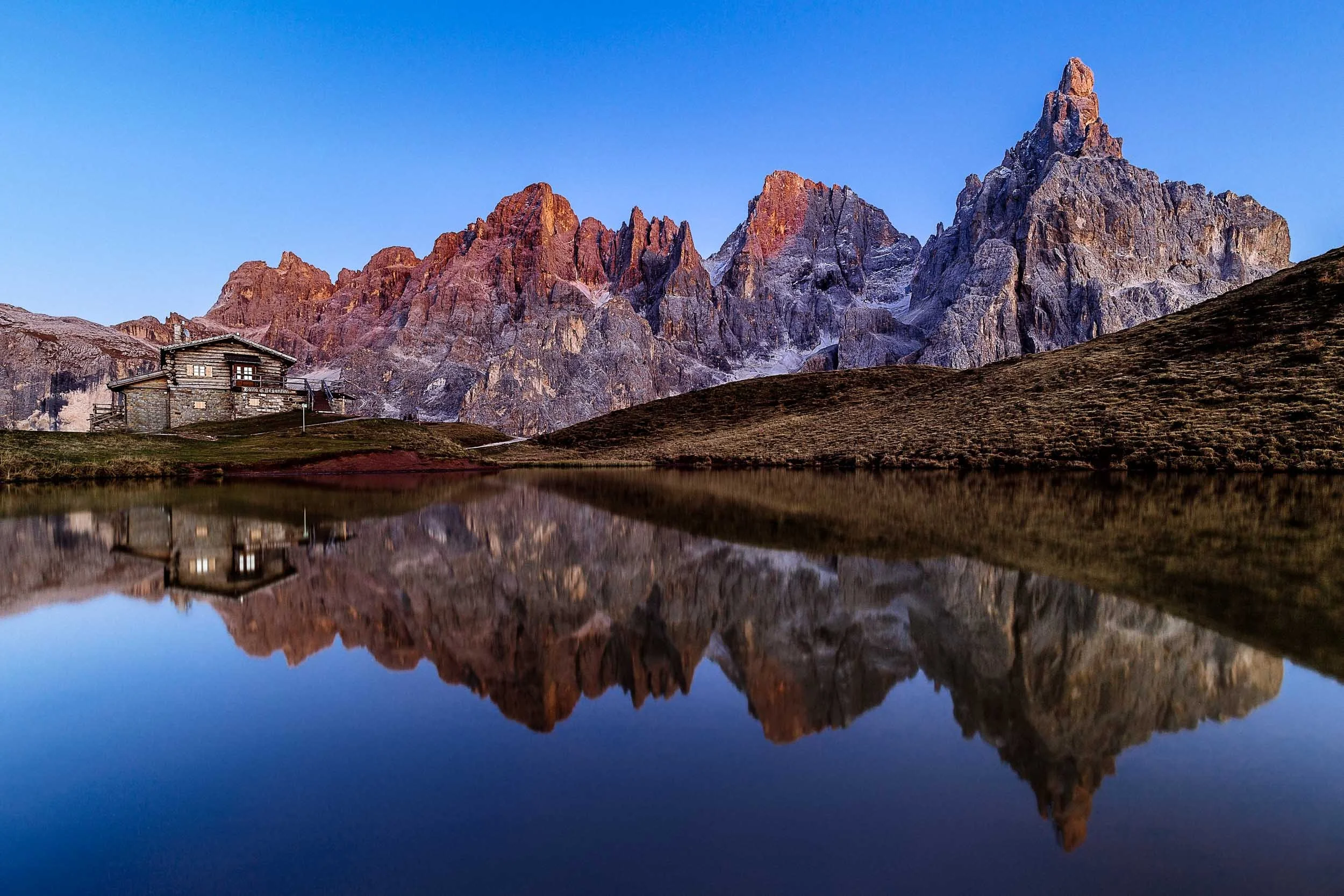 The alpine cabin Baita Segantini alongside the Pale di San Martino mountain range being mirrored in an alpine lake. 