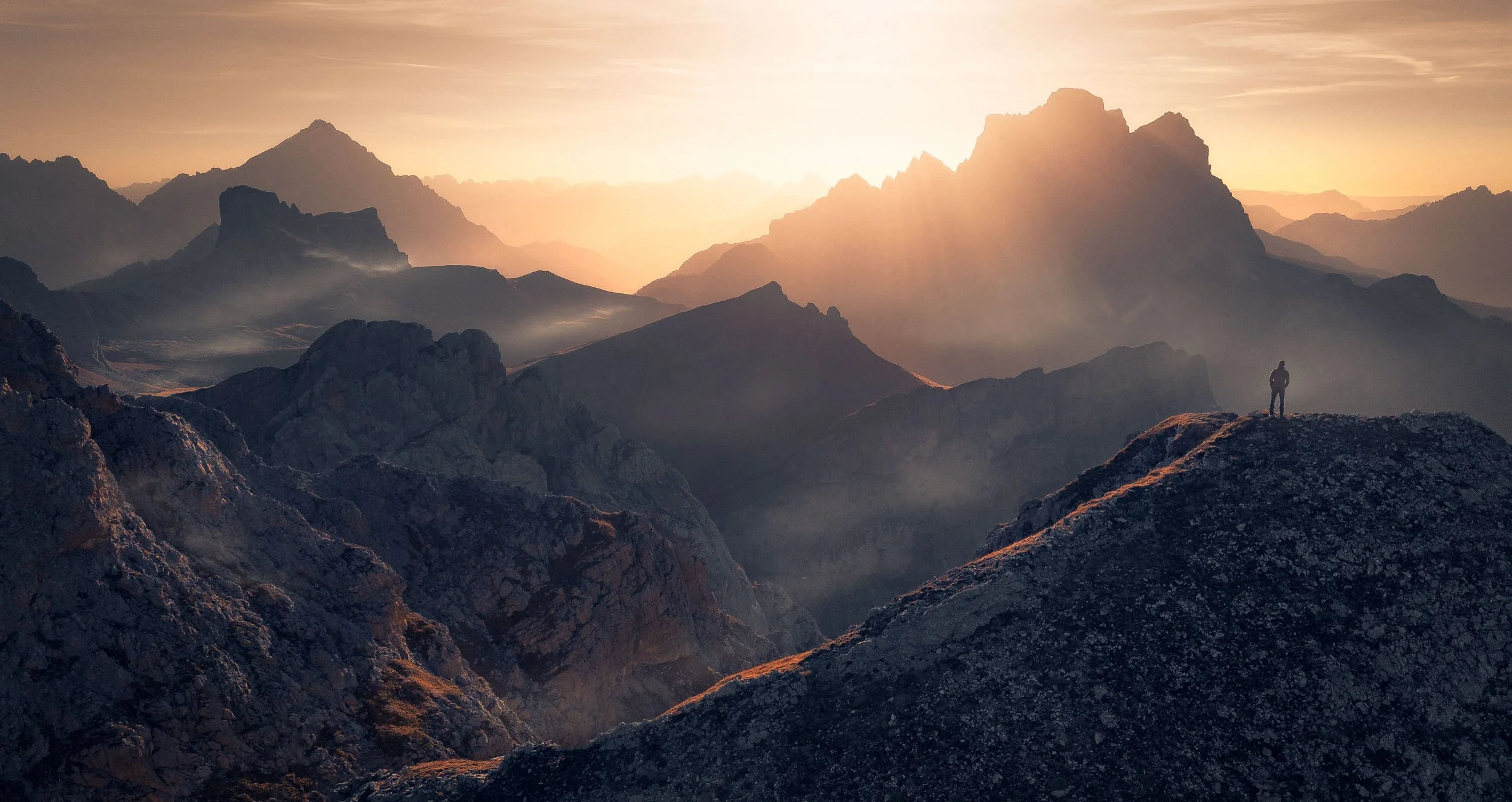 Hiker standing at the top of Monte Cernera watching the sunrise over Malga Mondeval in the Dolomites, Italy with Monte Pelmo and Antelao in the background. 