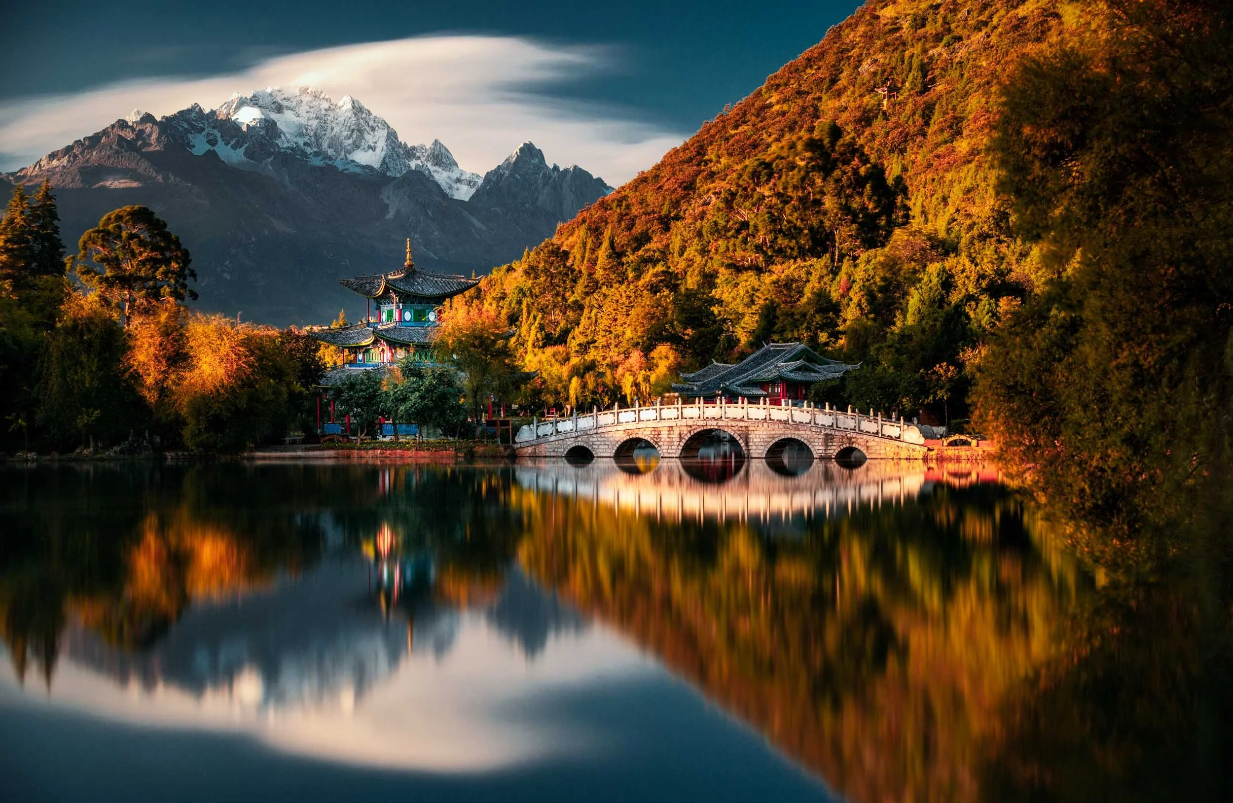 Reflections of the moon-embracing pavilion and a white marble bridge in the black dragon pool located in Jade Spring Park, at the foot of the Jade Dragon Snow Mountain in Lijiang, China. 

