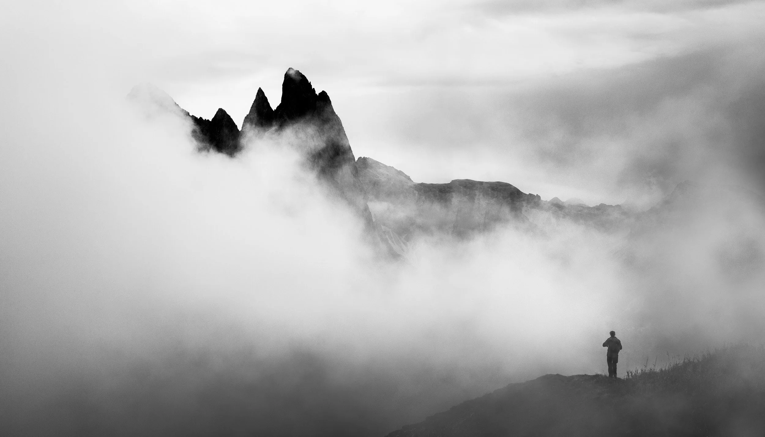 Man standing at the edge of a plateau taking a photo of the peaks of the Geisler mountain range of Seceda in the Dolomites, Italy, which are barely looking through a sea of clouds that rises from the valley. 