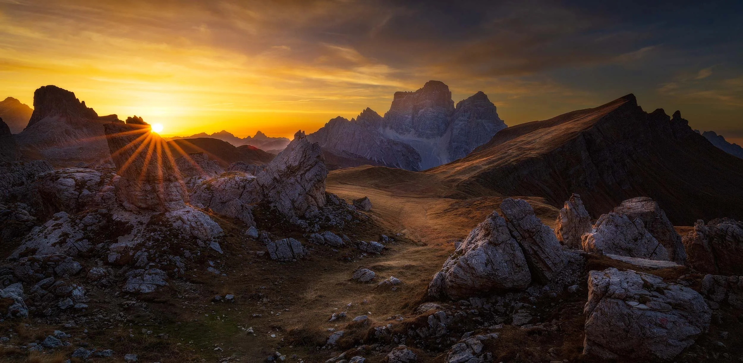 Malga Mondeval with its big boulders scattered in the valley during a warm autumn sunrise with Monte Pelmo towering in the background and the sun just peaking over the horizon showing a beautiful sun star.  